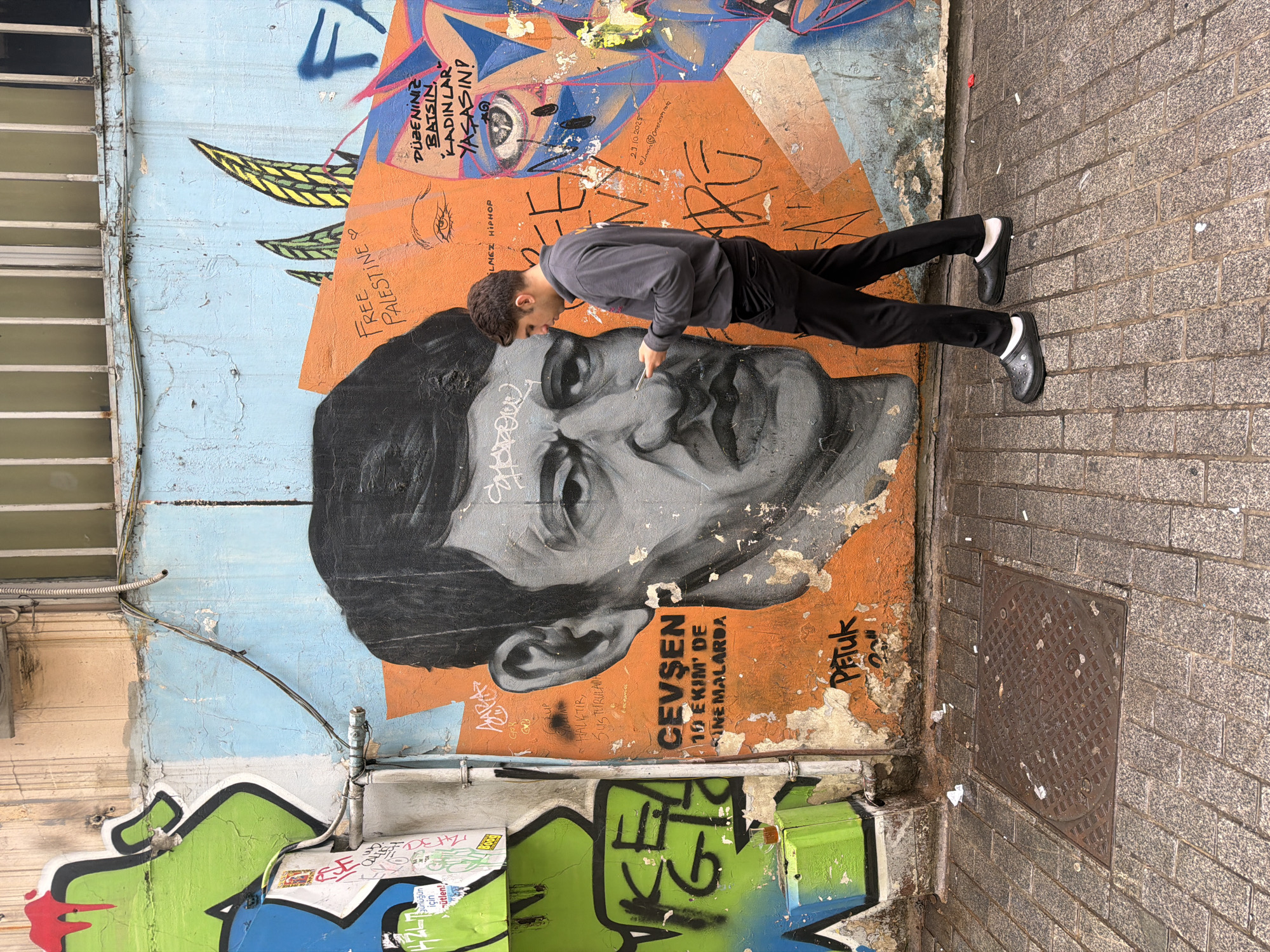 A man in black pants and a gray sweatshirt is walking along a cobblestone street in front of a graffiti-covered wall. He is looking down at his phone. The graffiti on the wall is colorful and abstract, with some recognizable images of faces and text. The wall is located in Kadıköy, Turkey.