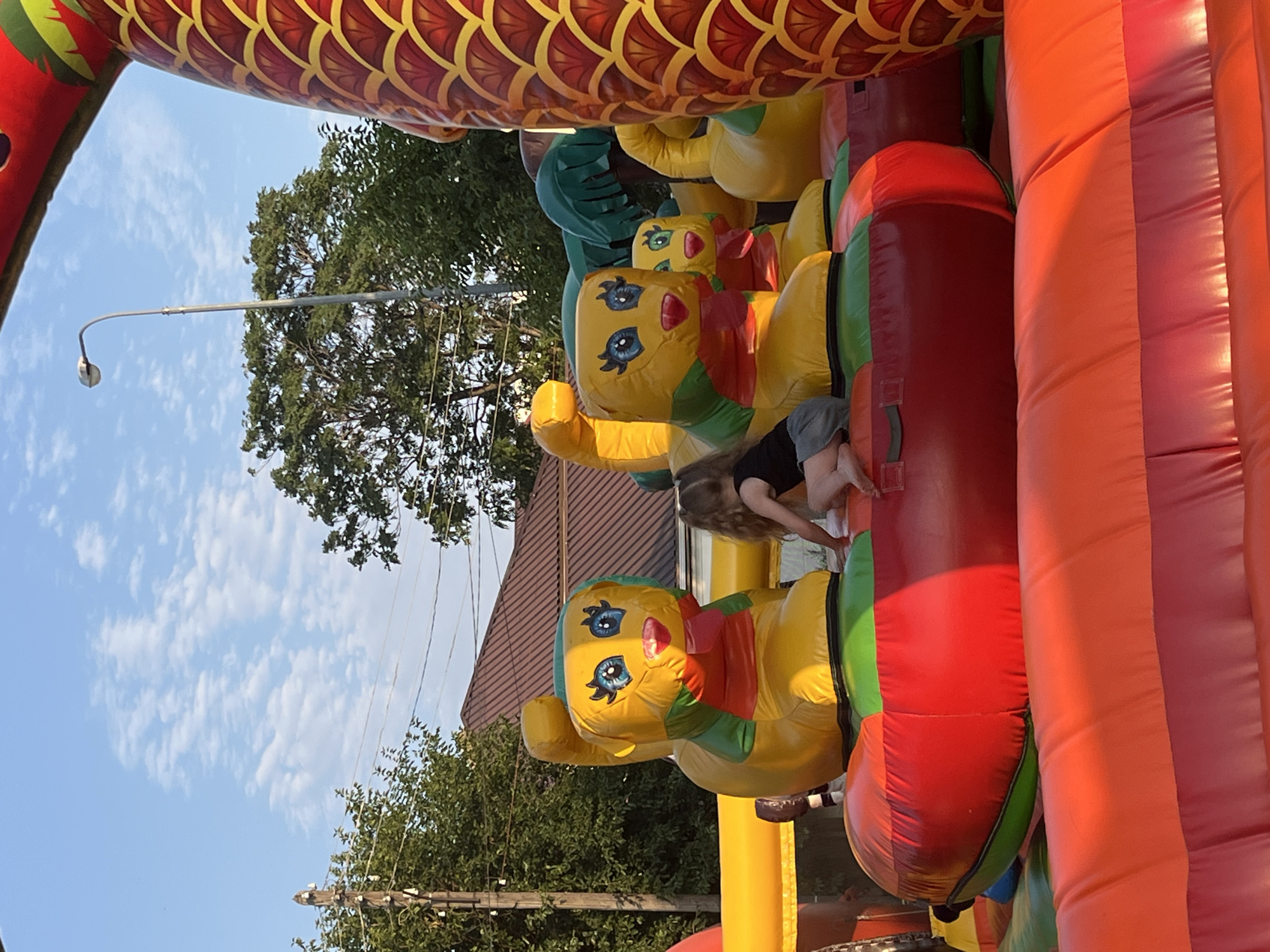 This is a daytime, medium-angle, eye-level outdoor shot of a bouncy castle attraction, likely in Astrakhan, Russia, given the style of housing and vegetation. The focus is on a section of the inflatable structure, which features brightly colored cartoon animal figures. In the foreground, a young girl with blonde hair, wearing a black tank top and grey shorts, is seated on the inflatable surface, looking down and to the right. Her right hand is extended downwards, and her left hand is resting on the inflatable. Behind her and to her left are two large, yellow inflatable cartoon ducks with blue eyes, red beaks, and green neck scarves. To the right of these ducks, a smaller inflatable duck is partially visible. The bouncy castle itself is a vibrant mix of red, orange, and green inflatable sections. In the background, a brown-tiled roof of a house is visible, along with green trees and a clear blue sky with scattered white clouds. A street lamp stands tall in the background, behind the bouncy castle and to the left. The overall impression is one of a children's entertainment area set up outdoors.