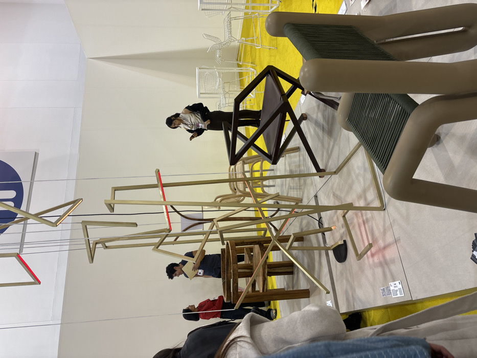 A showroom or exhibition displays various seating options. Several people are examining the furniture. A complex, linear art installation hangs in the foreground with integrated red light accents. Multiple chairs are visible, including a brown wooden folding chair, a beige chair with a woven green seat, and stacked wireframe chairs in the background. The flooring is a mix of yellow carpet and neutral-toned tile.