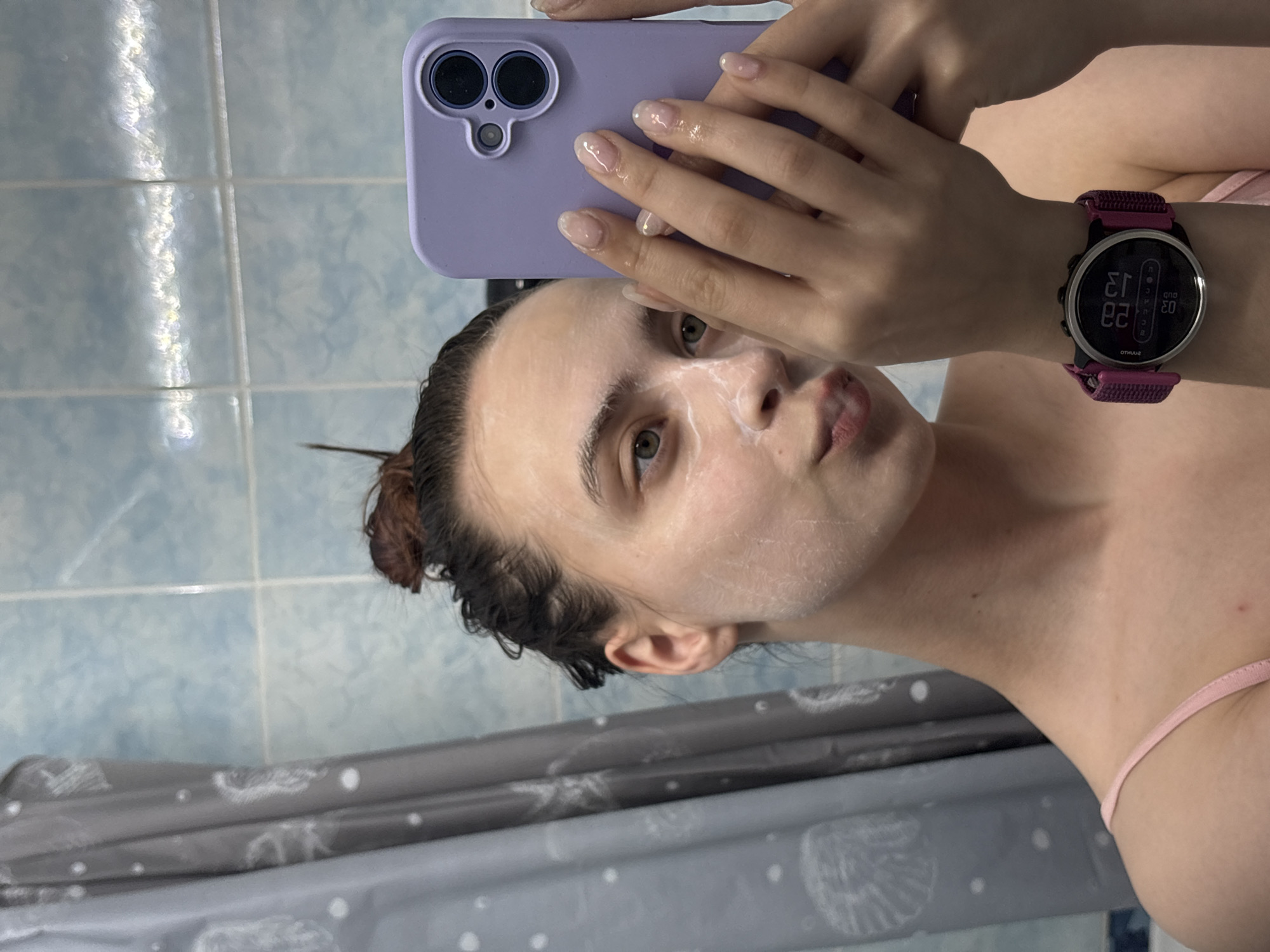 A selfie shows a woman with a white facial mask, holding a lavender-colored smartphone in her right hand to take the photo. The woman has a smartwatch on her left wrist, with the time displayed as approximately 12:23. She is in a bathroom, with light blue tiled walls visible in the background, along with a shower curtain.