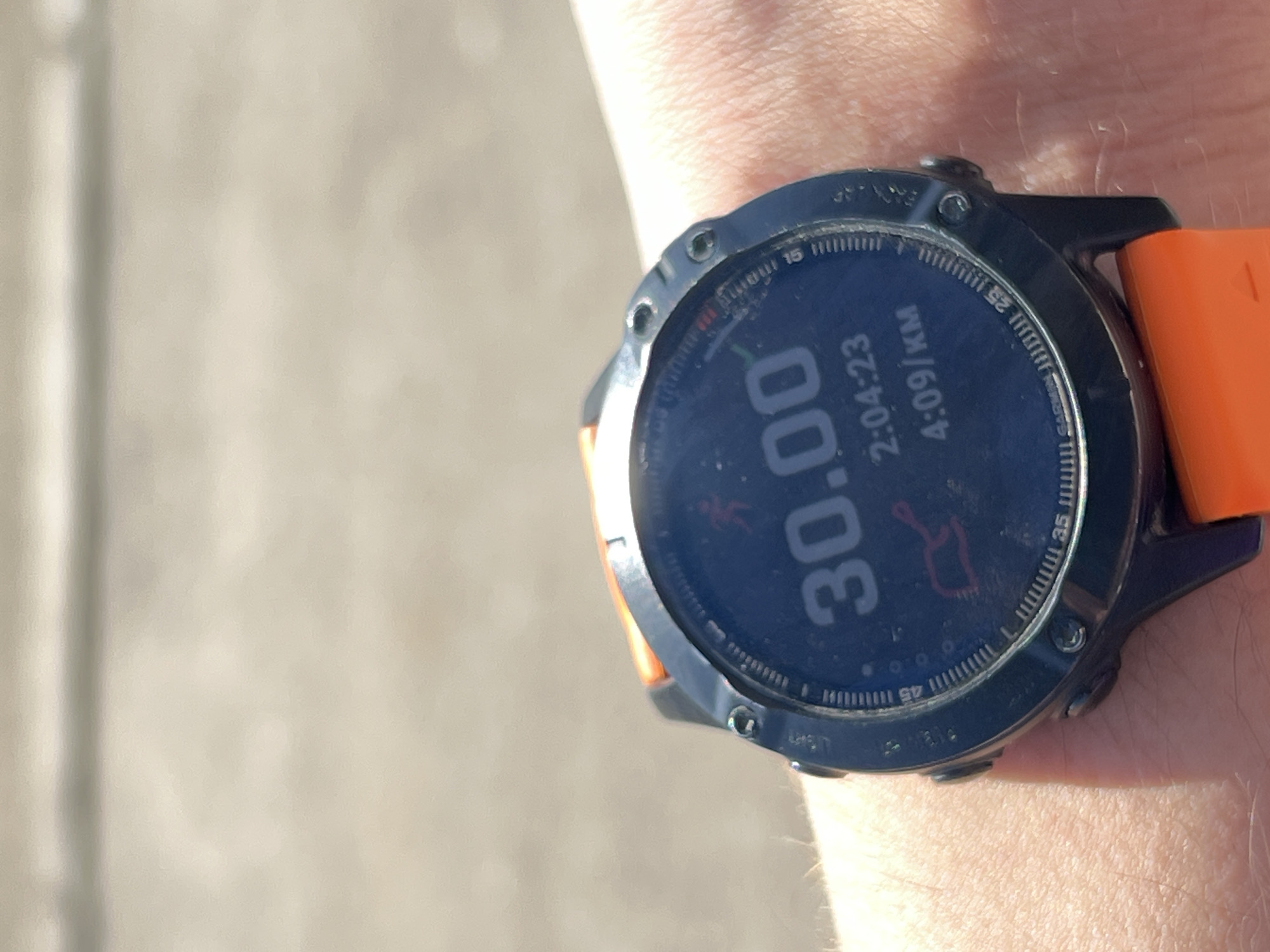 A close-up shot shows a wrist wearing a smartwatch with an orange band. The watch display shows the number "30.00", along with "2:04:23" and "4:09/км". The band is bright orange. The watch itself appears to be dark with a textured rim. The surrounding skin of the wrist is pale. The background appears blurry and out of focus, suggesting a street or outdoor environment. Kazan and Russia are not discernible from the image.