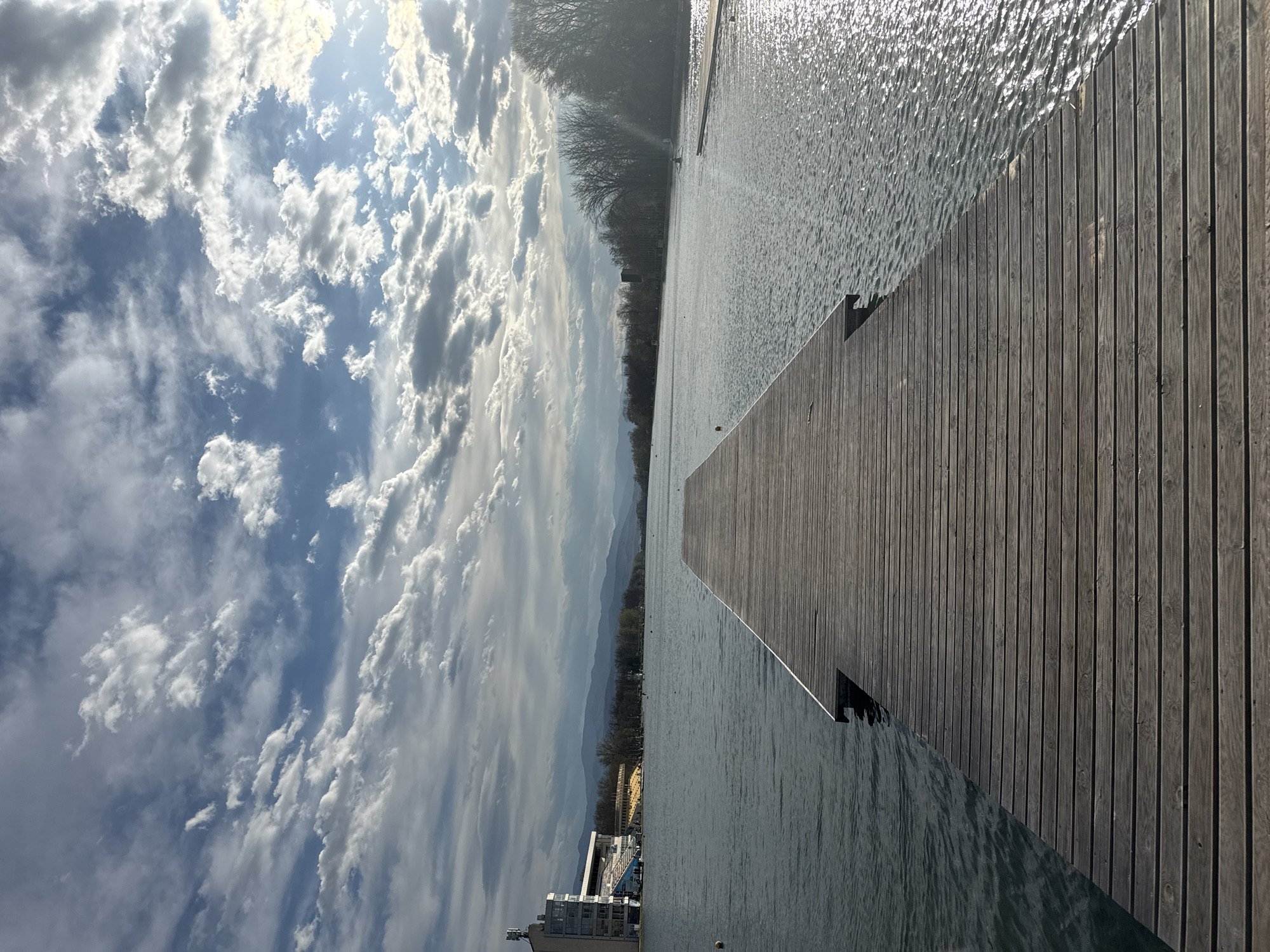 A ground-level, medium shot of a wooden dock extending into a body of water, likely Lake Plovdiv, in Plovdiv, Bulgaria. The weathered, gray-toned dock has vertical planks and juts out towards the lake. The water's surface reflects sunlight, creating a shimmering effect. The background displays a hazy skyline with trees and buildings along the shore, including structures that look like modern apartment complexes. Above, a cloudy sky with varying shades of blue and white adds a dramatic backdrop to the scene. The photo is oriented vertically.