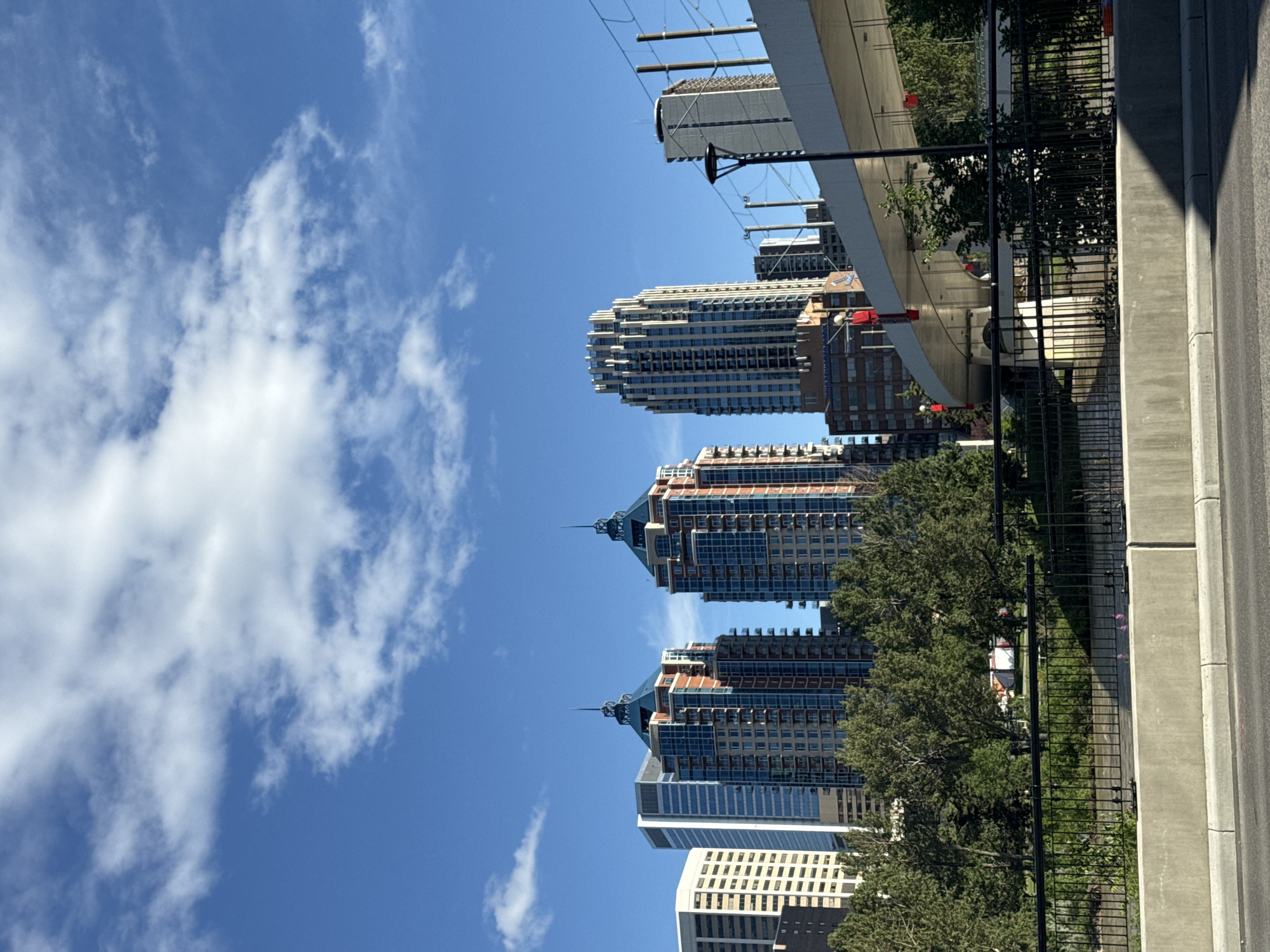 The image depicts a view of the Calgary skyline on a clear, sunny day with scattered white clouds. In the foreground, a concrete barrier and a black metal fence line a walkway or pathway. Beyond this, a body of water, likely the Bow River, is visible, with lush green trees on its banks.
Dominating the mid-ground and background are several tall buildings. Prominent among these are three distinct skyscrapers with pointed, pyramidal roofs, featuring a mix of glass, brick, and metal facades in shades of blue, orange, and beige. To the left of these, a modern, rectangular, glass-faced skyscraper stands out. Further to the right, another tall, fluted skyscraper with a pale facade is visible, along with a shorter, lighter-colored building with a round feature at its top.
A curved overpass or bridge structure is also visible, supported by columns and running diagonally across the image from right to left. This structure appears to be part of an urban transit system, possibly for light rail, with overhead wires and poles present. Red decorative elements are attached to the underside of the overpass.
The overall scene suggests an urban setting within Calgary, Canada, showcasing its modern architecture juxtaposed with natural elements like trees and water, under a bright blue sky. There are no visible people or animals in the image. The environment appears clean and well-maintained.