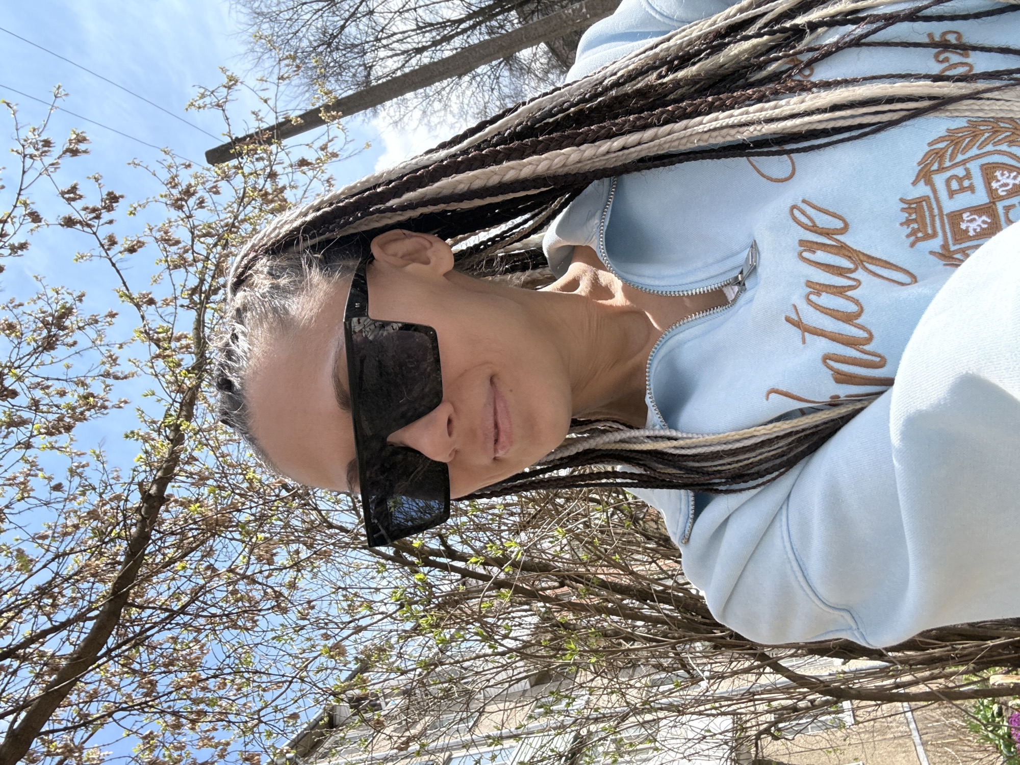 The image is a vertical outdoor selfie, featuring a person from the chest up, captured from a low angle looking upwards. The individual has long, multi-toned braided hair (dark brown, light brown, and off-white/grey) draped over their shoulder and chest. They are wearing large, rectangular dark sunglasses that cover a significant portion of their face, and a light blue zippered hoodie. A faint, subtle smile is visible on their lips beneath the sunglasses.
On the front of the light blue hoodie, there is embroidered text and a crest in a golden-brown color. The word "vintage" is clearly visible in a script font, and to its right, a crest-like emblem with design elements and possibly letters such as "P" and "R" or "B" is present.
The background consists of a bright, clear blue sky with some scattered clouds. Bare tree branches, some with small, nascent green leaves, frame the person against the sky, indicating springtime. A utility pole or similar wooden structure is partially visible in the upper right. Hints of buildings can be seen through the tree branches in the lower background. The overall lighting suggests it is daytime, likely a sunny morning or afternoon.