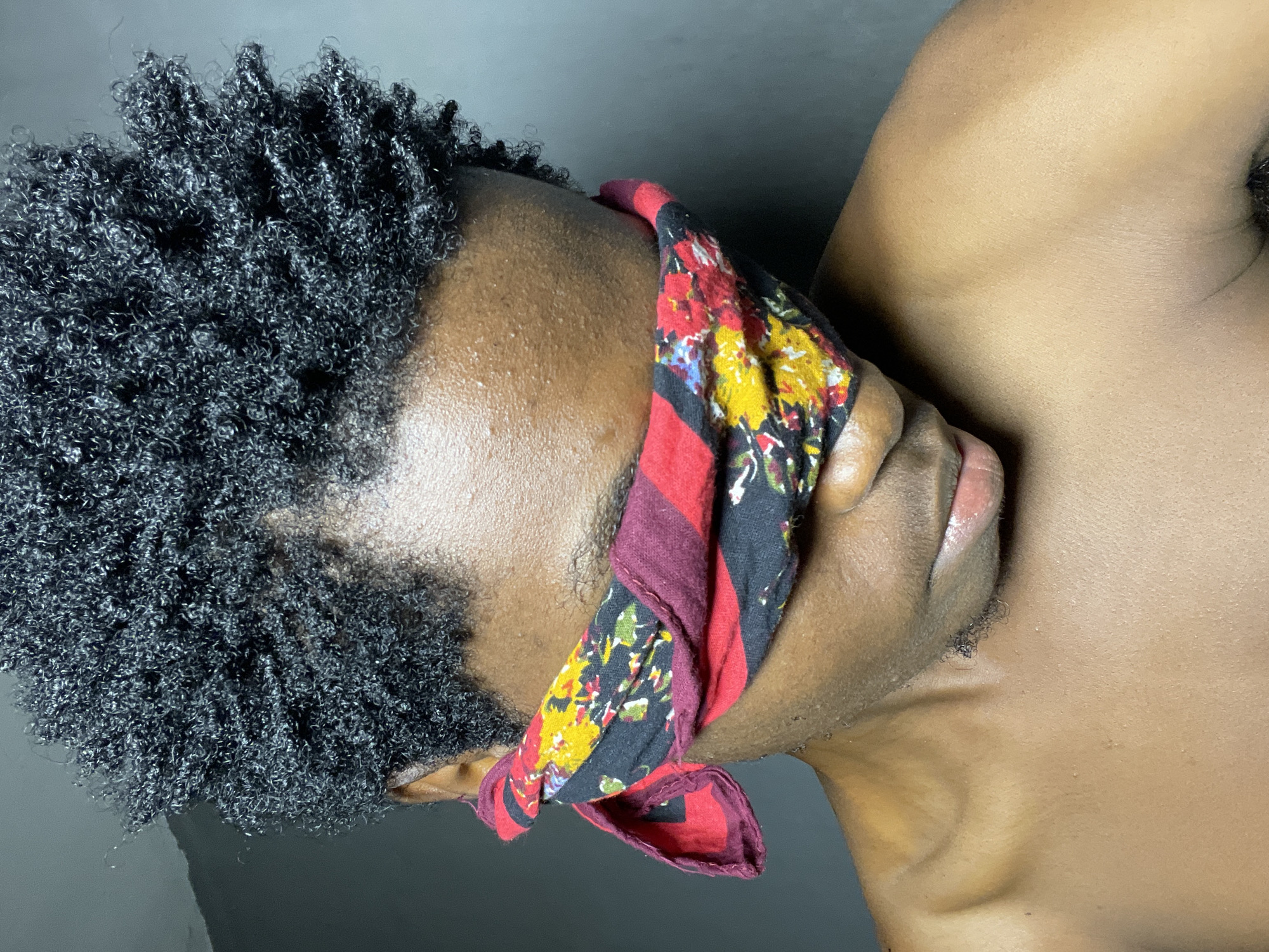 The image shows a close-up of a person's head and shoulders, with a colorful fabric blindfold covering their eyes. The person has dark, curly hair and brown skin. The background is a plain, dark gray wall. There is no information in the image that can be used to determine the location in the city of Nkpor, Nigeria.