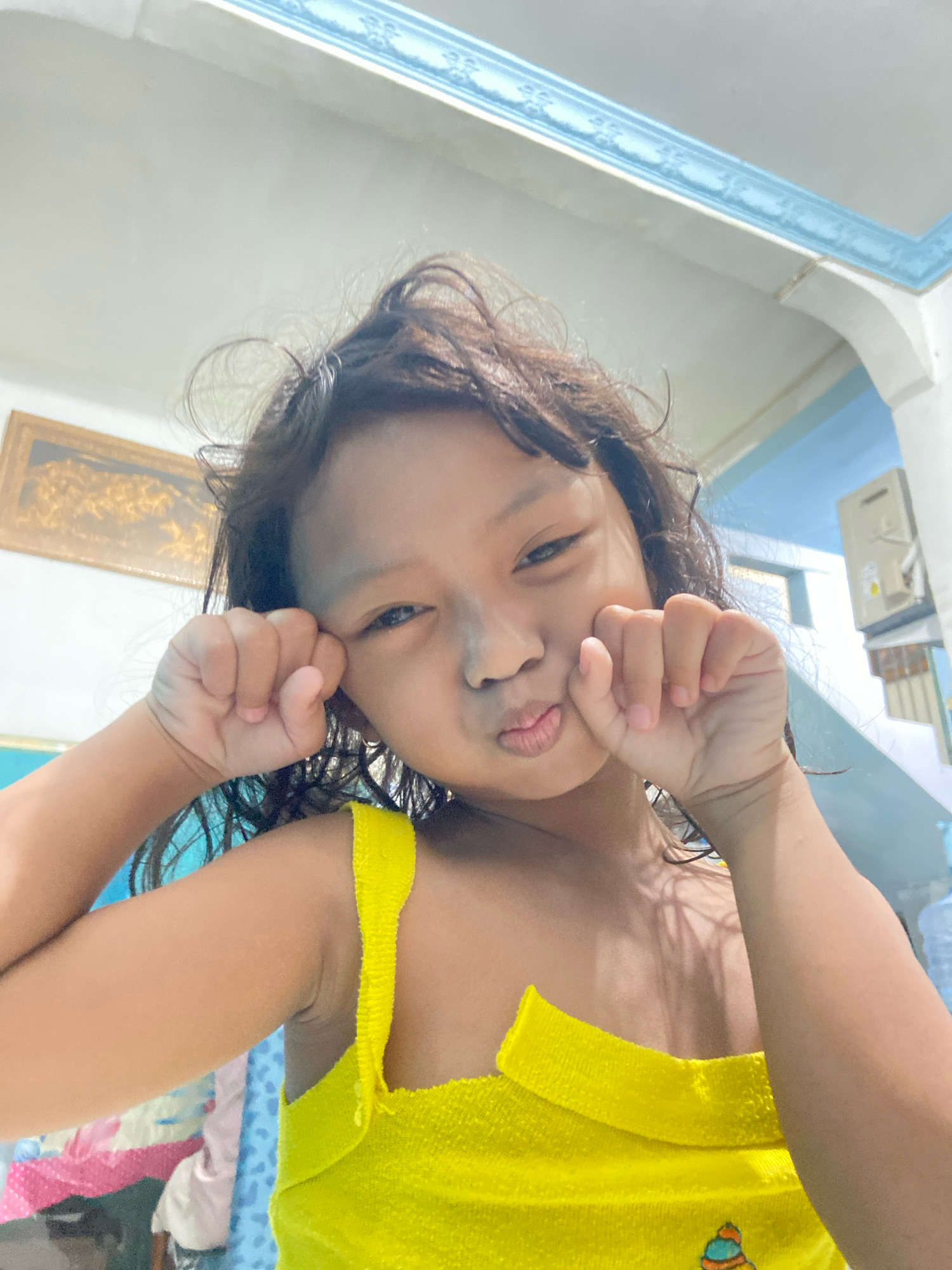 A young girl with dark, curly hair makes a "fish face" at the camera, holding her fists to her cheeks. She is wearing a bright yellow, terrycloth tank top. The background appears to be indoors, with a framed picture on the wall and a blue decorative trim near the ceiling. The lighting suggests it is daytime. The exact location in Gelam, Indonesia, is not discernible from the image.