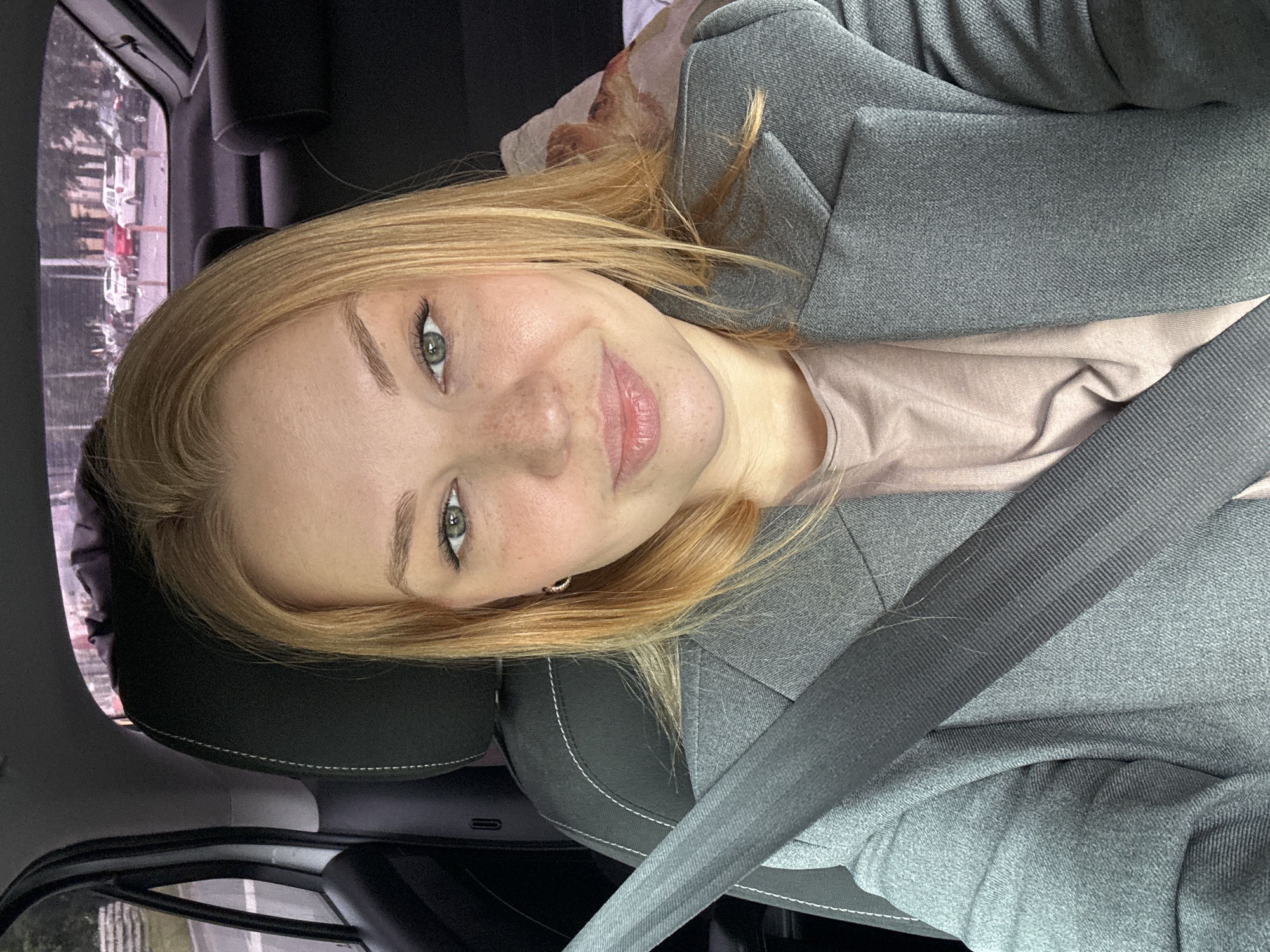 A blonde woman with green eyes and freckles is taking a selfie in the passenger seat of a car. She is wearing a grey blazer over a pink shirt, and a grey seatbelt is visible across her chest. The interior of the car, including the headrest of the driver's seat, is visible. Through the car windows, a street with parked cars and buildings can be seen. The scene appears to be in an urban environment, possibly Zhukovo, Russia, given the context.