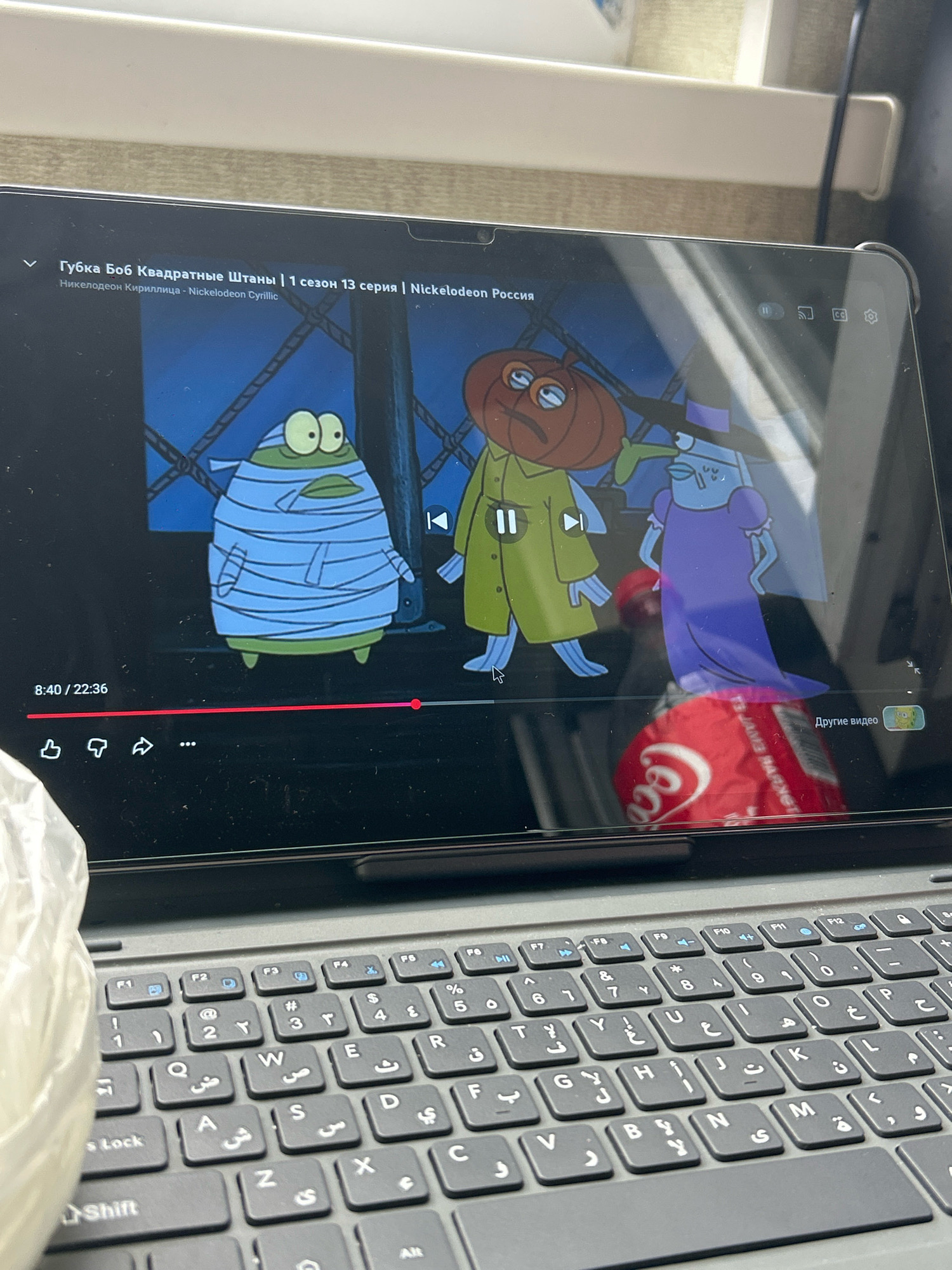 A tablet computer displays the animated show "SpongeBob SquarePants" with Russian subtitles. The screen shows three characters: a mummy-like creature on the left, a character with a pumpkin head in the middle, and a tall, slender character in a purple dress on the right. The playback progress bar indicates the video is at 8:40 of 22:36. Below the screen, a portion of a black keyboard with Arabic and Latin letters is visible. The tablet is placed on a surface that appears to be a desk or table. The background is out of focus and shows a light-colored wall and some blurry objects. The scene suggests a casual setting, possibly at home or a workspace. No specific location in Khyrdalan, Azerbaijan is discernible from the image.