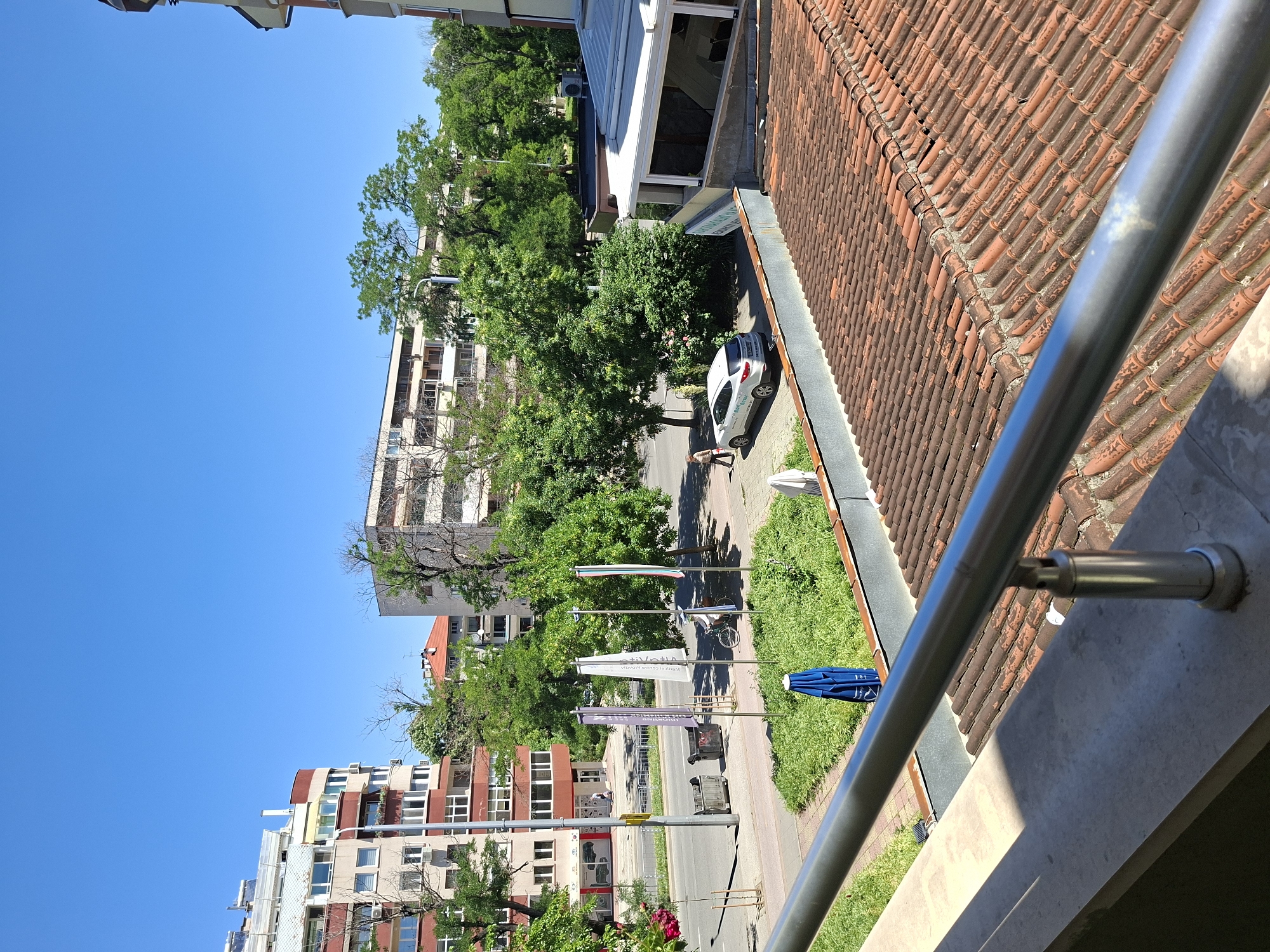 The image shows a street scene in Plovdiv, Bulgaria, taken from an elevated position, possibly a balcony. Buildings line the street, with a mix of residential and commercial structures visible. The sky is clear and blue. Trees provide a green canopy over parts of the street. Several flags are displayed along the sidewalk. A parked car and a pedestrian can be seen in the vicinity of one of the buildings. The balcony railing is visible in the foreground, and red tiled roofs are present in the midground.