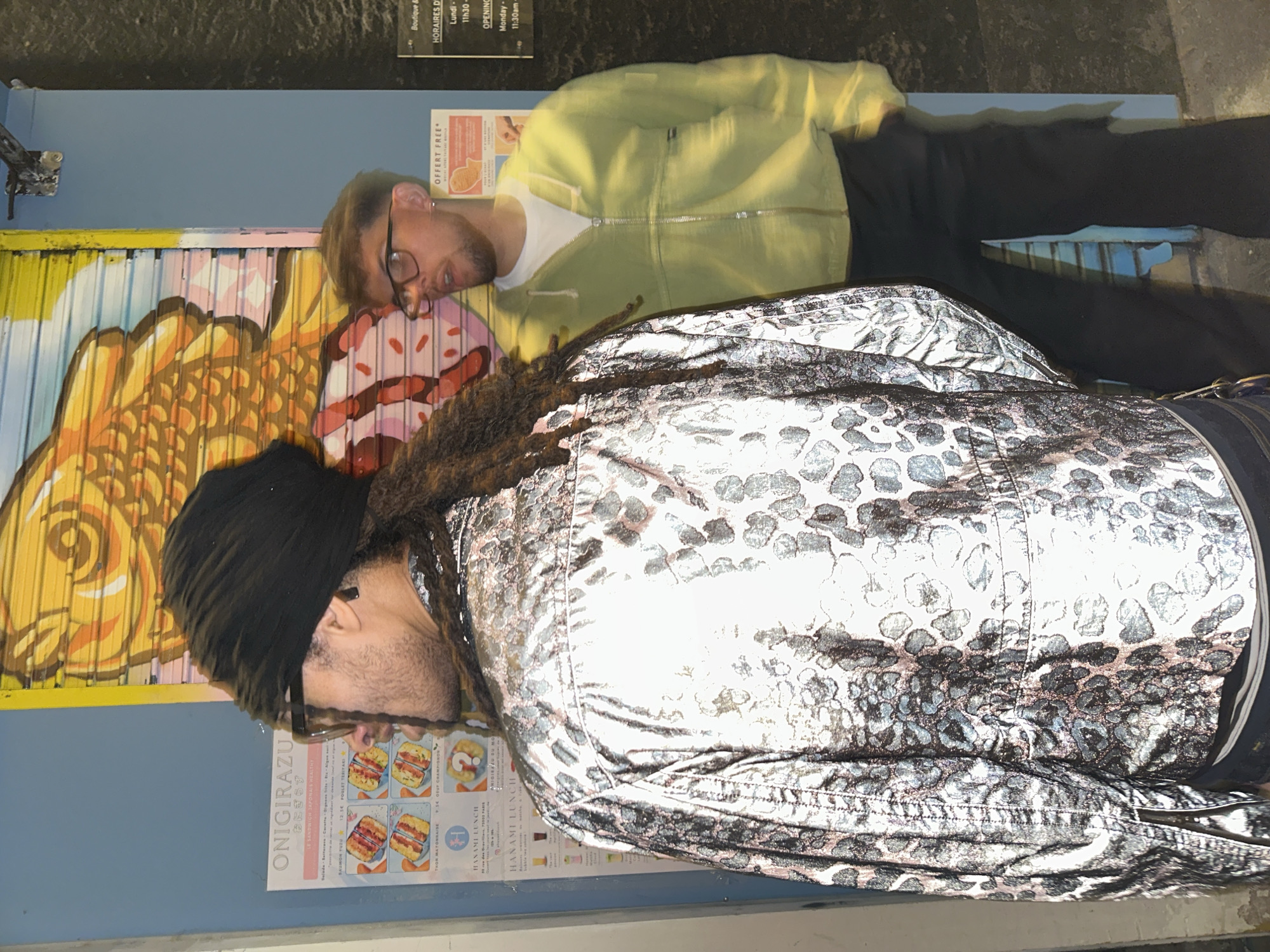 The image displays two men standing outdoors in what appears to be an urban setting, likely Paris, France, as indicated by visible text. The background features a vibrant, multi-colored mural on a wall to the left, depicting abstract or stylized forms in yellows, oranges, pinks, and blues, including a large fish-like shape and a striped, cake-like object. To the right of the mural, the wall is a solid blue, with several posters attached. The ground beneath the men is dark and textured.
In the foreground, on the right side of the frame, a man with dark dreadlocks and a black beanie wears a distinctive silver or metallic jacket featuring a dark, mottled, possibly leopard-print pattern. He also has a beard and glasses, and his head is turned downwards. Behind him and slightly to his left stands another man wearing a light yellow or lime green zip-up hoodie over a white t-shirt, black pants, and glasses. He also has a beard and is looking downwards.
Visible text on the posters includes "ONIGIRAZU" (Japanese sandwiches), "HANAMI LUNCH," and menu items such as "POULET TERIYAKI" and "SAUMON FUMÉ." Opening hours are listed as "Lundi-" / "Monday-" from "11h30" / "11:30am." The address "75002 PARIS" confirms the location in Paris, France. "OFFERT FREE" is also noted on one poster. The scene appears to be set during the daytime, possibly in front of a restaurant or shop. The men are not engaged in any specific activity beyond standing and looking.