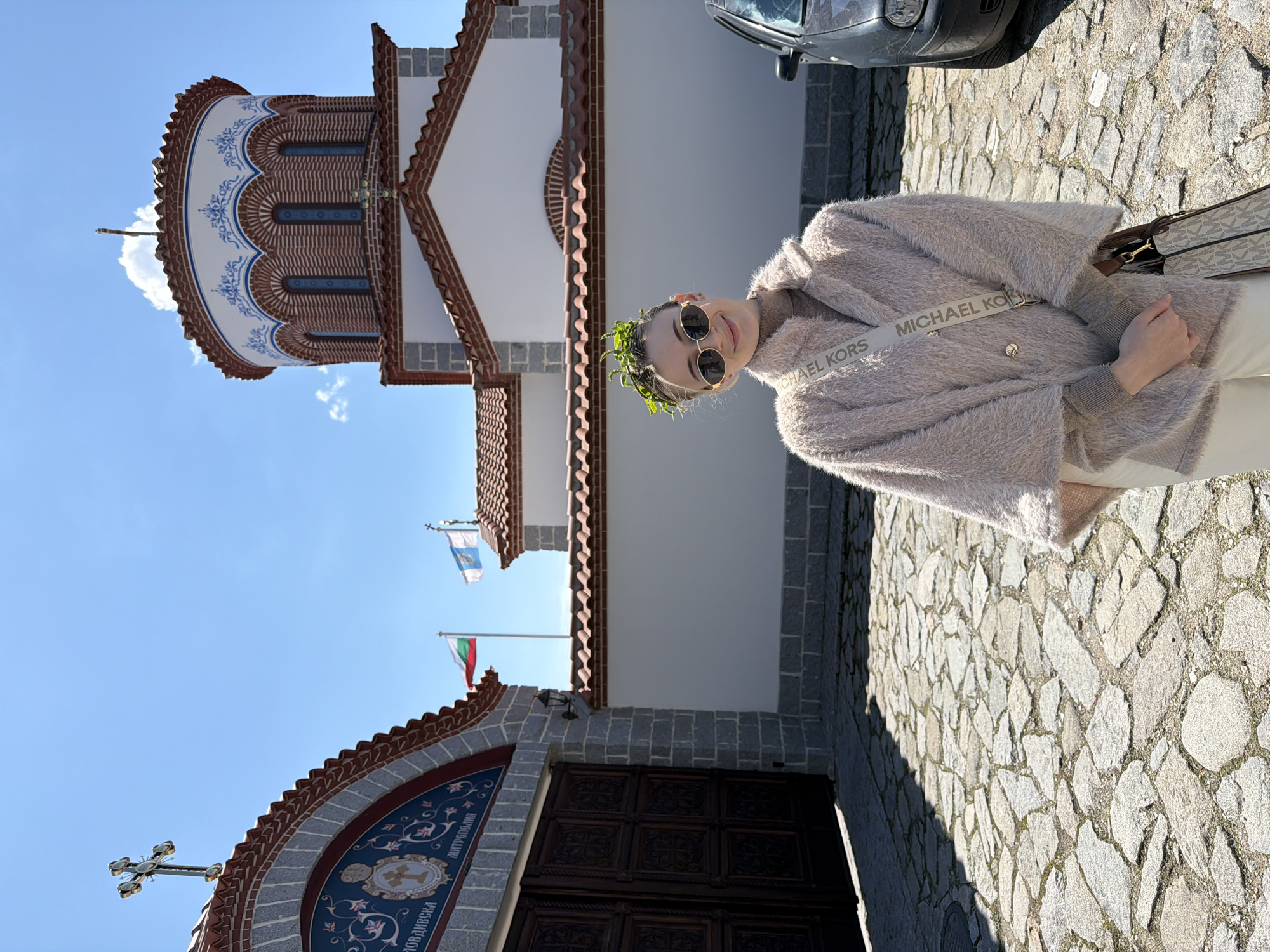 A medium shot depicts a woman with dark hair and fair skin, wearing round sunglasses and a garland of greenery, standing on a cobblestone surface in front of a church. The church is painted white with brick detailing, featuring a tower with a blue and white design. Flags of Bulgaria and the city of Plovdiv flank the church entrance. The woman is wearing a fluffy beige jacket with a Michael Kors shoulder strap. A dark-colored car is visible in the upper right corner. The sky is clear and blue. The image appears to be taken in Plovdiv, Bulgaria.