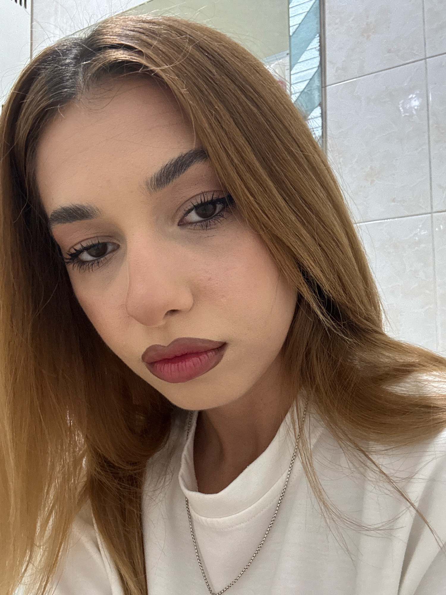 A close-up, head-and-shoulders selfie of a young woman with light brown hair and brown eyes. She is wearing a white t-shirt and a silver chain necklace. Her lips are painted with a dark reddish-brown lipstick. The background appears to be a tiled wall, possibly in a bathroom, with a reflection of striped blinds in a mirror. The image does not contain any information indicating the location is the city of Getahovit, Armenia.
