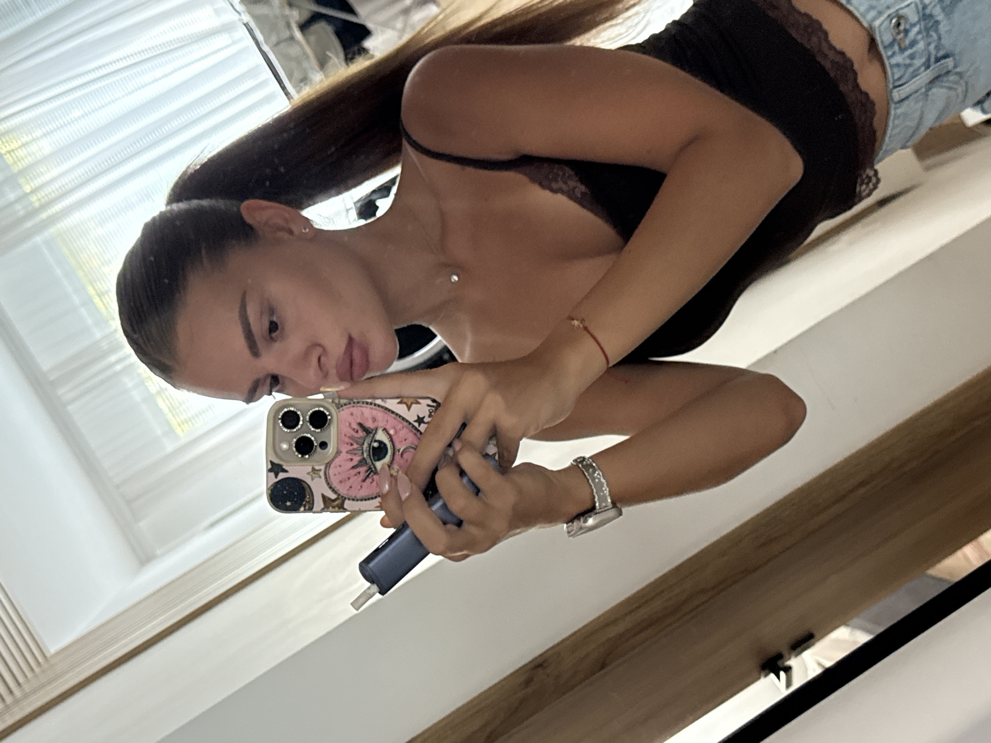 In Plovdiv, Bulgaria, a young woman with long, dark brown hair styled in a high ponytail takes a mirror selfie. She is wearing a black lace-trimmed camisole and light blue denim jeans. In her right hand, she holds a blue electronic device, possibly a vape pen, and in her left, she holds a smartphone with a pink phone case featuring a large eye design with lashes and stars. She is also wearing a silver watch with a studded band on her left wrist and a thin silver necklace with a small pendant. A red string bracelet is visible on her right wrist. The reflection shows a brightly lit room with sheer white curtains in the background and a wooden doorframe to her left.