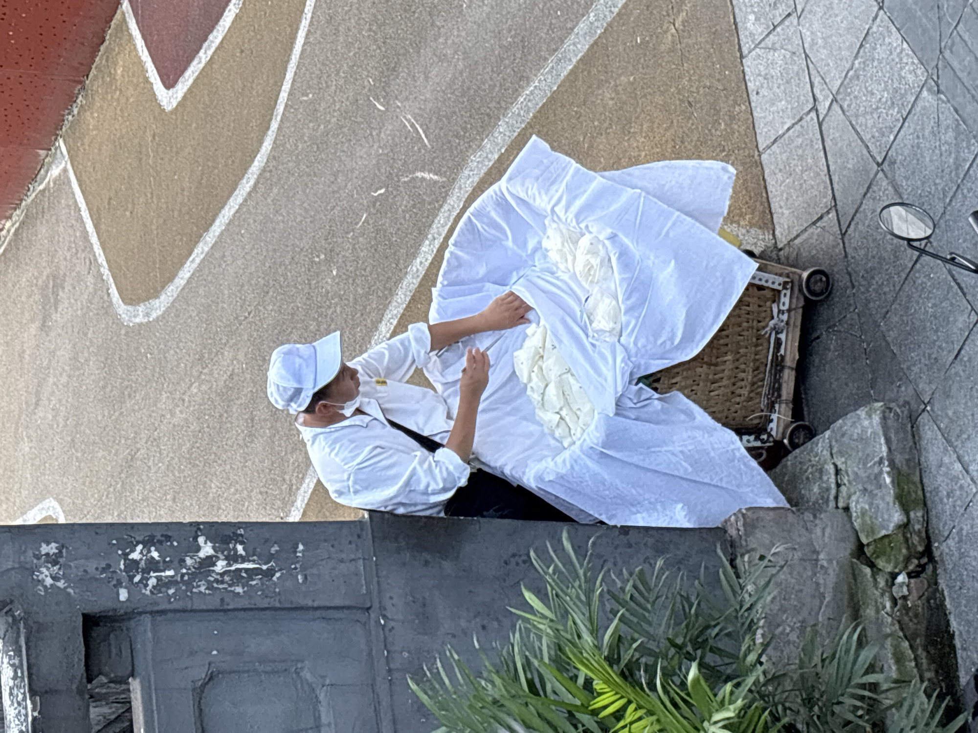 An overhead shot captures a person in a white long-sleeved shirt and white cap tending to a large, woven basket filled with white items, covered with a white cloth, positioned on a wheeled cart. The person is wearing a light blue face mask. The basket is situated on a paved area with a pattern of square stones, adjacent to a darker gray, textured surface that appears to be a wall or column. To the left, lush green foliage is visible. The background shows a tan and reddish-brown surface with white painted lines, suggestive of an outdoor recreational or traffic area. The scene is outdoors, under natural light.