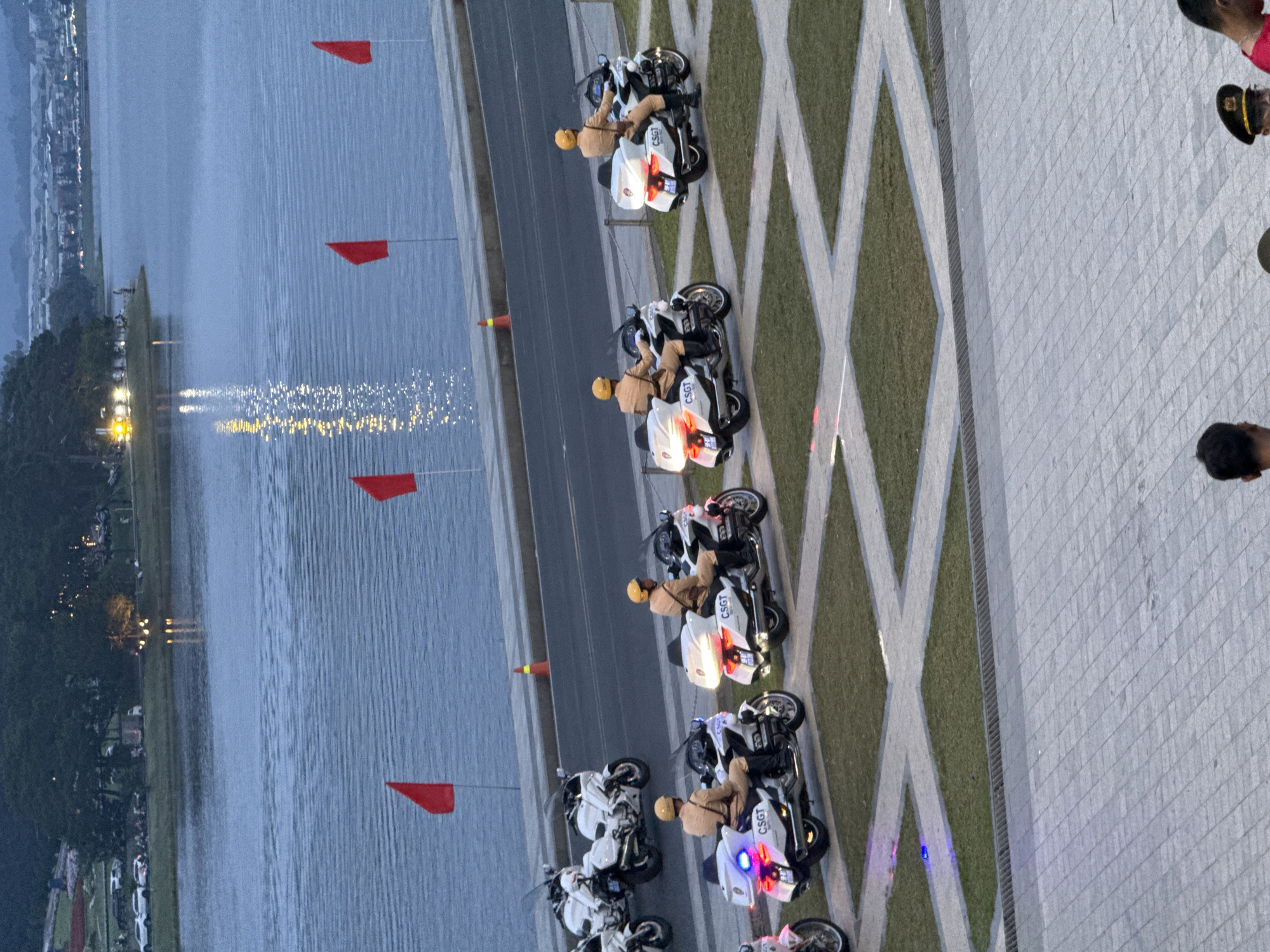 The image depicts a serene scene along a body of water, likely Xuan Huong Lake in Da Lat, Vietnam. In the foreground, a line of motorcyclists in uniform, presumably traffic police, are positioned on a paved area adjacent to a road. Their white motorcycles are equipped with police lights, some of which are illuminated with blue and red hues. The riders are wearing light brown uniforms and yellow helmets. Beyond the road, red flags are visible, standing tall against the calm water. The water reflects the faint lights from the distant cityscape and trees lining the shore. In the background, the Da Lat landscape is hinted at with lush greenery and a hazy skyline, characteristic of the city's mountainous terrain and diffused light. The overall atmosphere is one of order and tranquility, with the uniformed riders suggesting a presence of authority or a planned event.