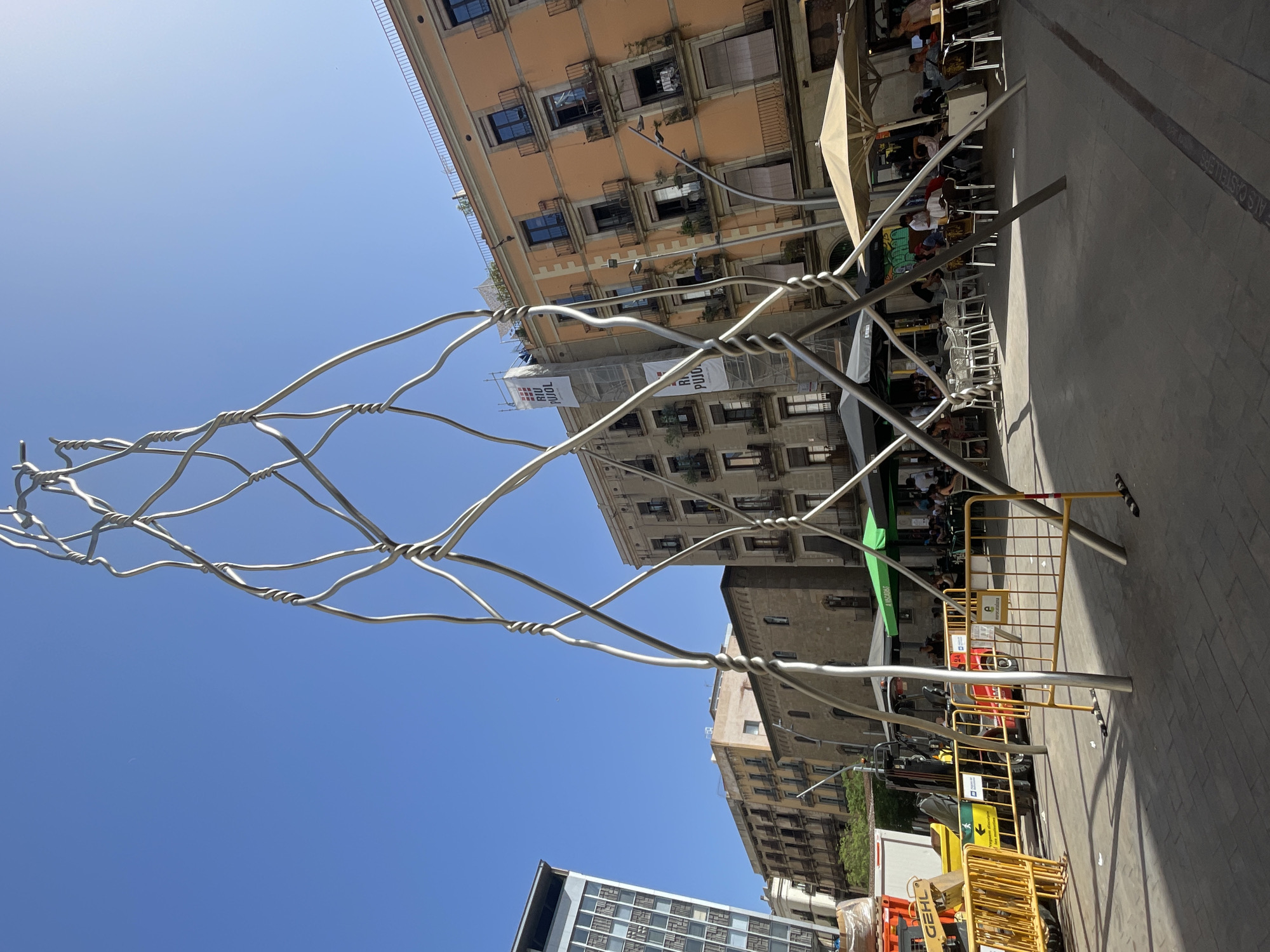The image captures an outdoor scene in what appears to be a city. The focus is on a large, abstract sculpture made of twisted metal wires, which dominates the foreground and extends upwards into a clear blue sky. The sculpture is silver and has a tangled, organic form.
In the background, there are buildings, some of which are residential with balconies and windows. One building has a banner with Spanish text that appears to say "RIU PUJOL". There are also some areas of scaffolding, suggesting ongoing construction or maintenance.
Further back, people are seated at tables under umbrellas, indicating an outdoor cafe or restaurant area. There are also some construction vehicles, including a yellow forklift and a white truck, along with yellow safety barriers. The ground is paved with what looks like cobblestones or patterned tiles.
The time of day appears to be daytime, with bright sunlight casting shadows. The sky is clear and blue, suggesting fair weather. The overall impression is of an urban environment that is possibly undergoing some development or has an artistic installation. The presence of people enjoying an outdoor meal suggests a relatively relaxed atmosphere despite the construction elements.