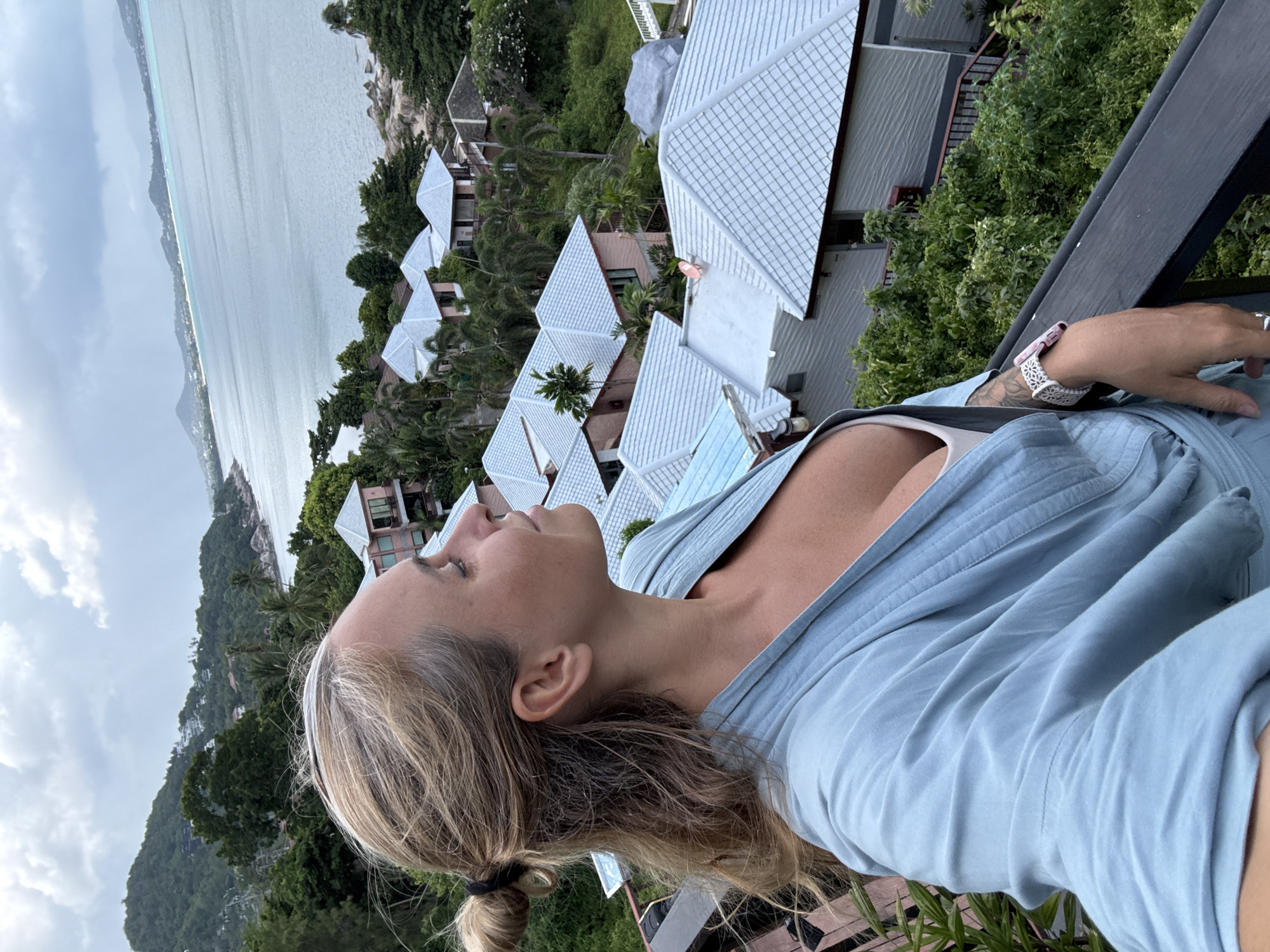 A medium shot captures a woman in profile, looking out over a landscape in Ko Samui District, Thailand. She has blonde hair pulled up in a bun and is wearing a light blue robe. Her left arm rests on a dark railing, and she wears a white watch with a pink band and a ring on her left hand. The background reveals a cluster of houses with light-colored roofs nestled amongst lush green trees. Beyond the houses, a body of water stretches out to a distant, hazy coastline under a cloudy sky. The perspective suggests the photo was taken from a balcony or elevated vantage point, offering a panoramic view of the tropical scenery.