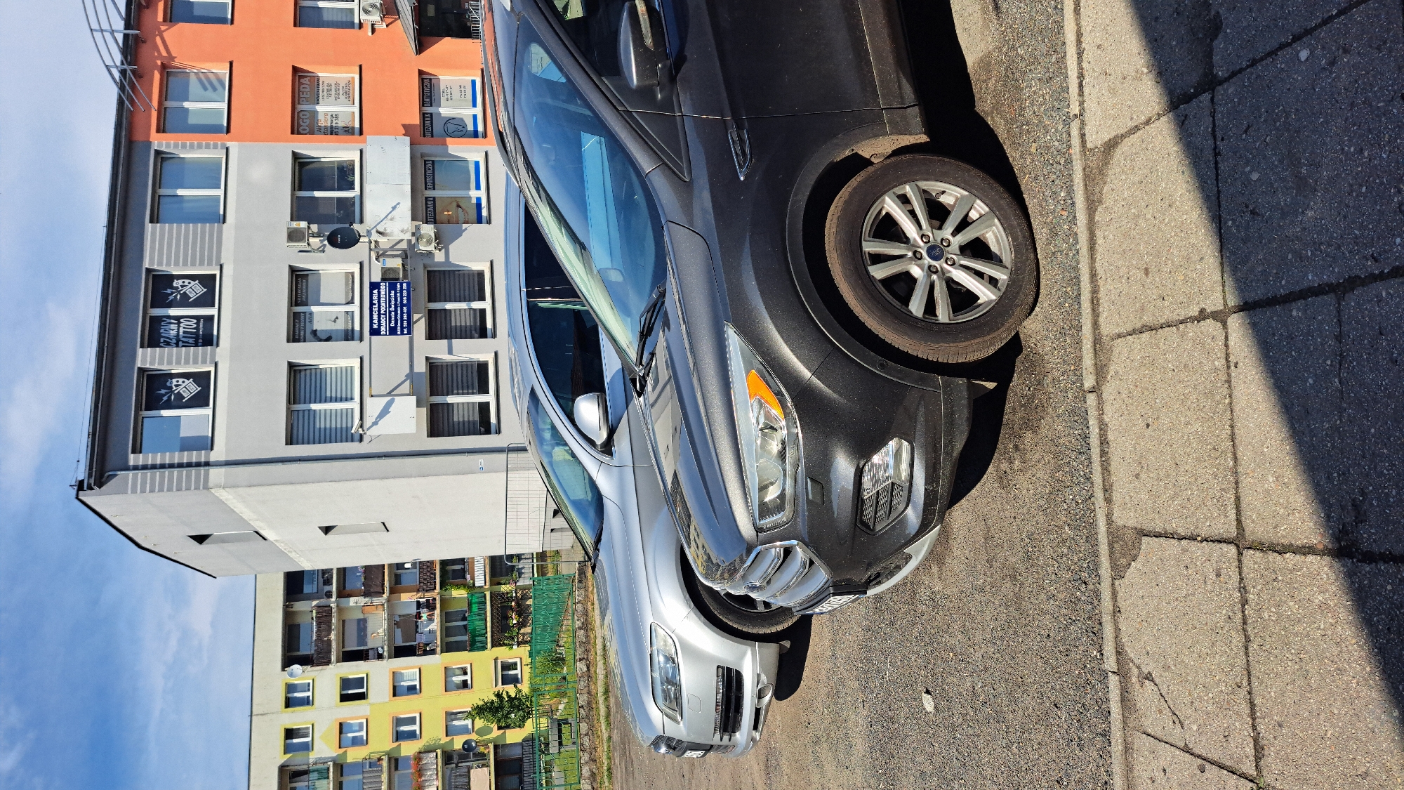 The image shows a street scene in Krosno Odrzańskie, Poland, on a sunny day. Two cars are parked on the right side of the image, with a sidewalk made of concrete slabs to their right. The cars are a silver SUV in the foreground and a dark gray SUV behind it. The SUVs are parked close to each other. In the background, there is a multi-story building with orange and gray facades. The building has many windows, and some of them display signage for businesses, including "CZARNY TATTOO" and "ROTEZOWNIA DENTYSTYCZNE." The building's architecture suggests it is a residential or mixed-use property. The sky is clear and blue, indicating fair weather. The lighting suggests it is daytime, likely mid-morning or afternoon, given the shadows cast by the cars and the building.