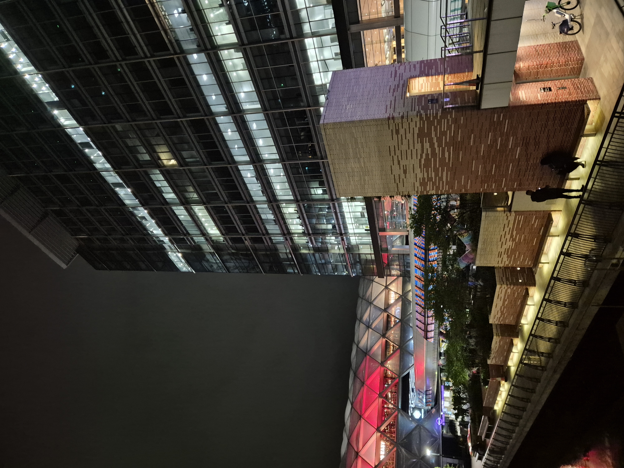 The image shows an urban night scene in London, United Kingdom. A tall, modern glass building dominates the left side of the frame, with numerous illuminated windows. Adjacent to this, a building with a textured brick facade rises. A pedestrian walkway with brick structures and benches runs parallel to a river or canal, where two individuals are visible. A bicycle is parked on the walkway. Further down, there is a building with a diamond-patterned glass structure illuminated with red and white lights. Between the buildings, trees are faintly visible, and there are subtle lights suggesting street activity. The sky appears dark and cloudy, suggesting an overcast night.