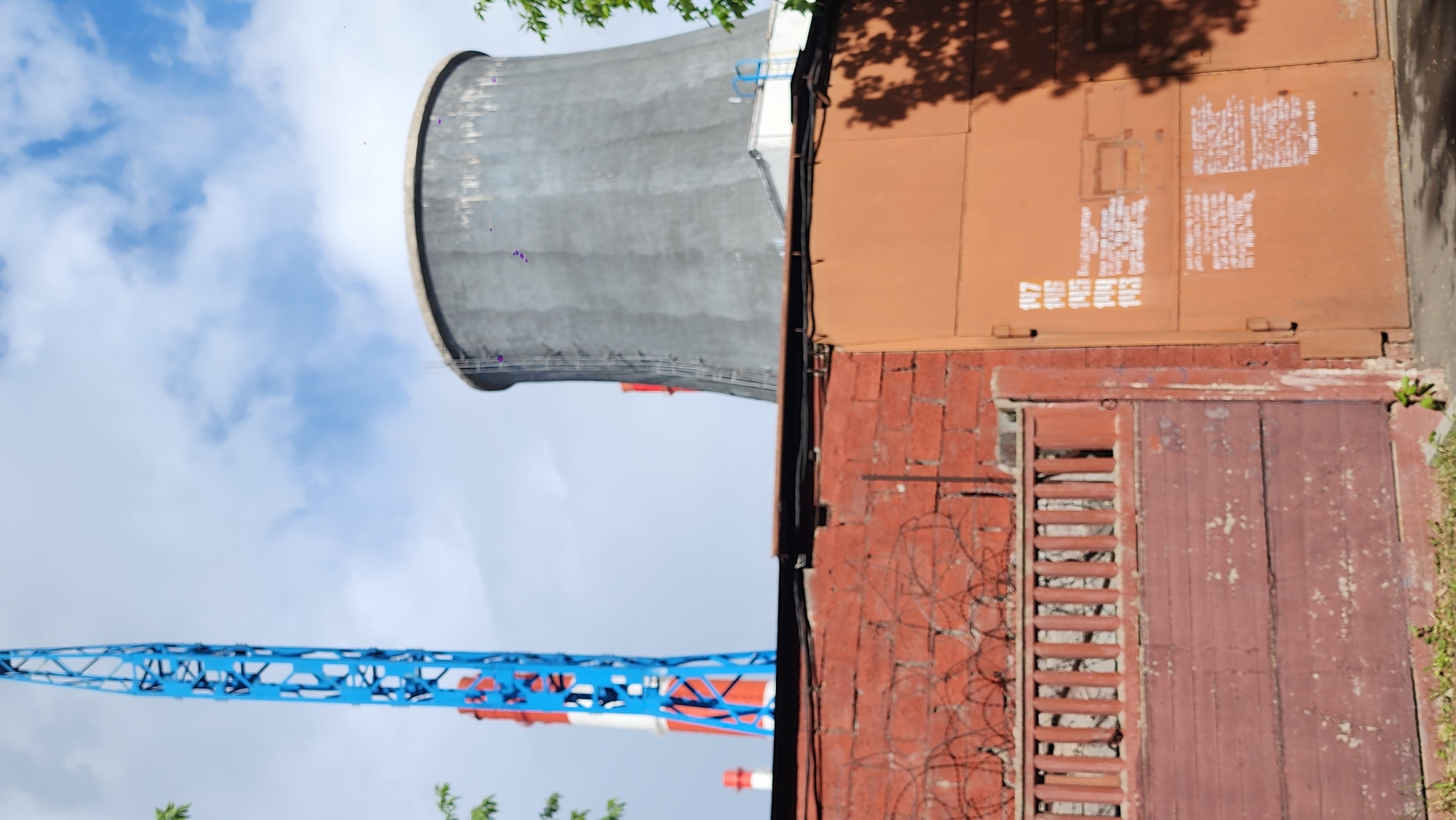 The image presents a landscape featuring a combination of industrial and infrastructural elements under a partly cloudy sky. A large, gray hyperbolic cooling tower, typical of power plants or industrial facilities, dominates the scene. To its right, a building with a multi-layered construction is visible, including brickwork and metal sections. A portion of a blue and red construction crane extends across the lower left area. The building's exterior displays details such as windows, a door, and barbed wire. Based on the elements in the image, it appears to be in Moscow, Russia.