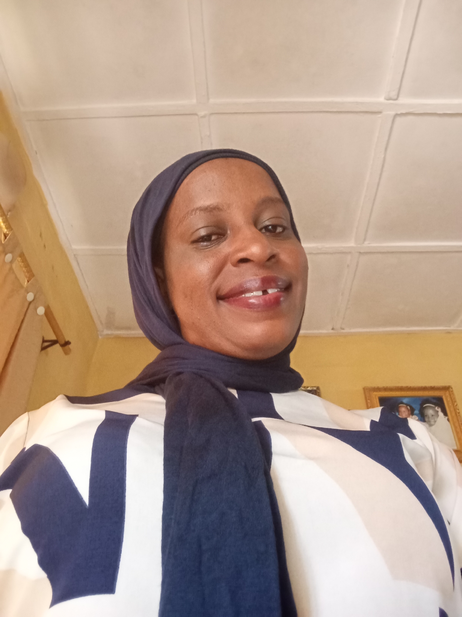 A woman wearing a navy blue hijab and a white and navy blue patterned blouse stands indoors, likely in a residence. The background includes a yellow wall, a white ceiling with panels, and framed pictures hanging on the wall. The woman is smiling and looking directly at the camera. The location is Narayi, Nigeria.