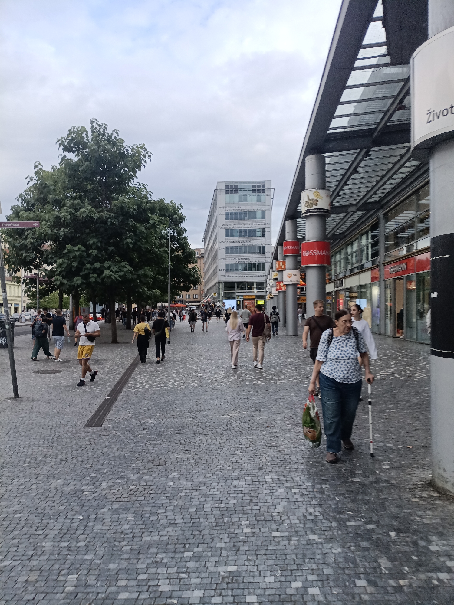 The image depicts a street scene in Prague, Czechia, likely in an urban commercial area. A cobblestone street dominates the foreground and midground, with a modern building complex featuring a glass roofed canopy on the right side. Several people are walking along the street in various directions. On the right, a woman with a cane and a shopping bag walks towards the viewer. A group of people are seen further down the street, some walking and some standing. Trees are present on the left side of the street, and a tall modern building with visible text on its facade is in the background. Signs with text like "ROSSMANN" and "Život" are visible on the buildings. The sky is overcast, suggesting a cloudy day.