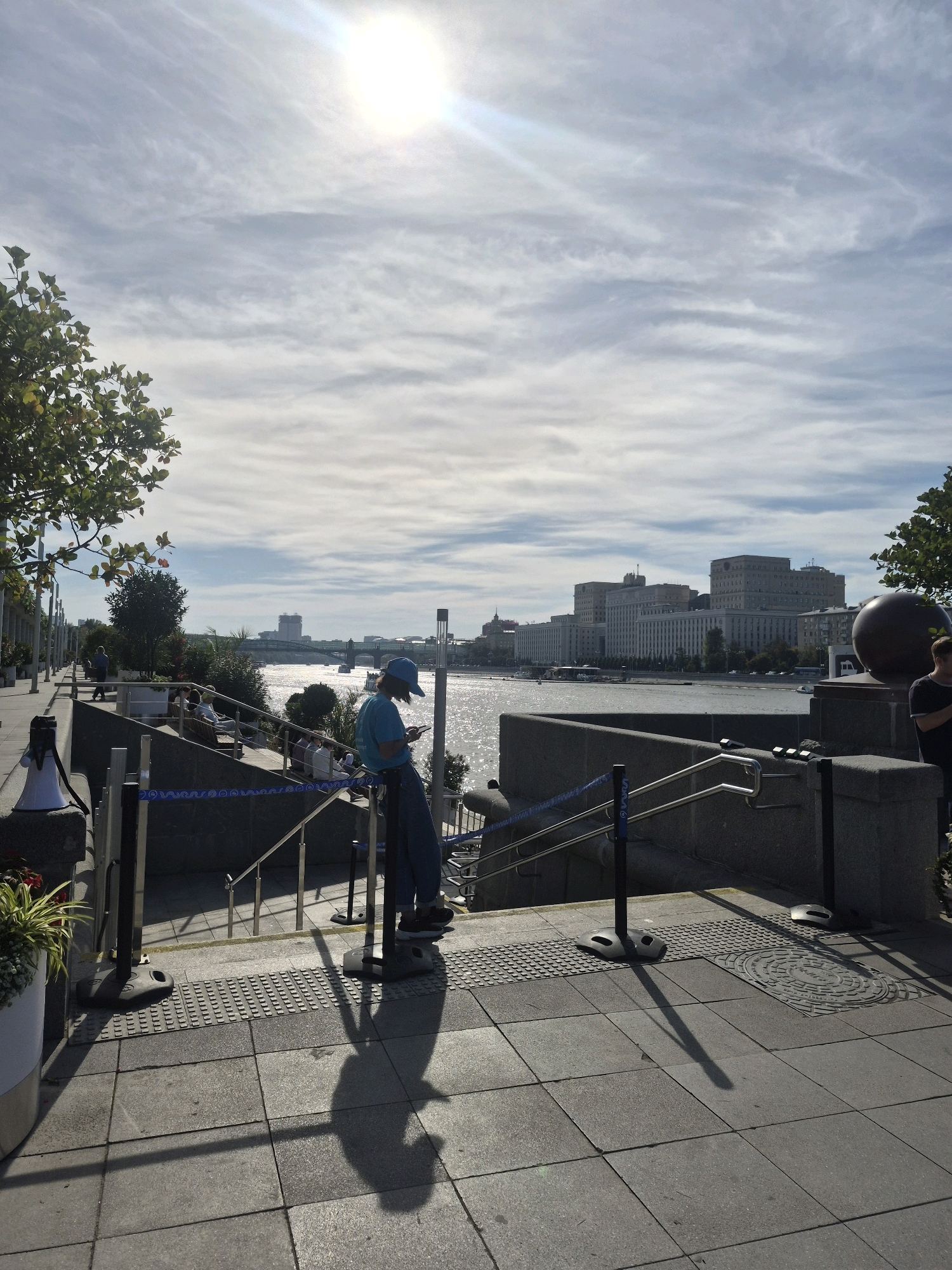 In an urban outdoor setting, a person wearing a blue cap and a blue t-shirt stands on a stepped walkway, looking down at their phone. They are leaning against a metal railing. Several other individuals are seated on benches further up the walkway, facing the water. Across a body of water, a large complex of buildings is visible. The sun is bright in the sky, with some clouds present. The walkway is paved with stone tiles, and there are planters with greenery on the left side of the image. Metal stanchions with blue decorative rope are also present, delineating the walkway. The overall scene suggests a public area, possibly a waterfront promenade.