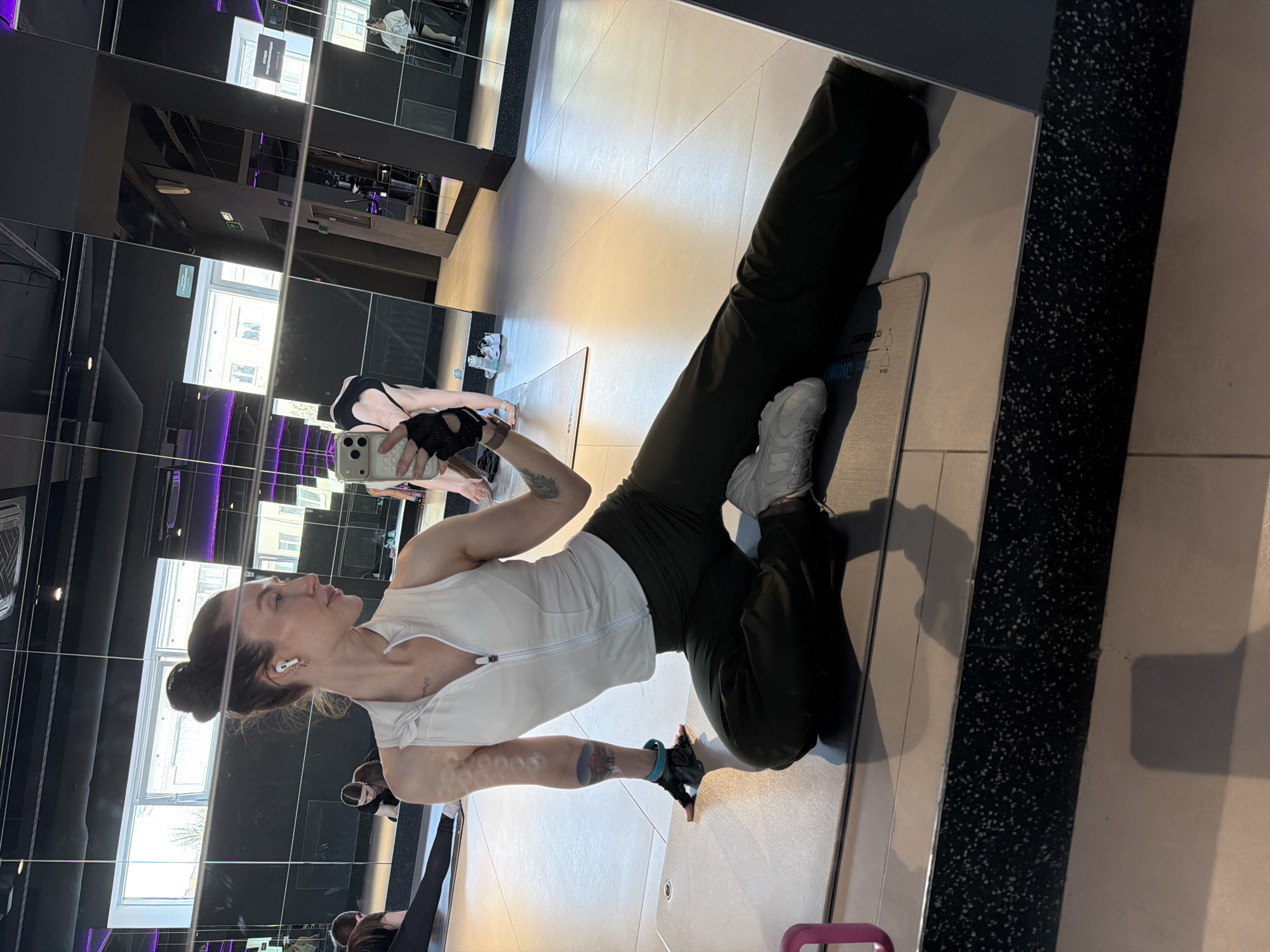 A woman is taking a selfie in a mirrored gym. She is wearing a white tank top, dark athletic pants, and light grey sneakers. She has her hair in a bun and is wearing earbuds. The mirror reflects the gym setting with exercise equipment and windows showing an urban scene, possibly in Perm, Russia. She is holding a smartphone in her left hand, wearing a black glove with a blue wristband. Her right knee is on a grey exercise mat.