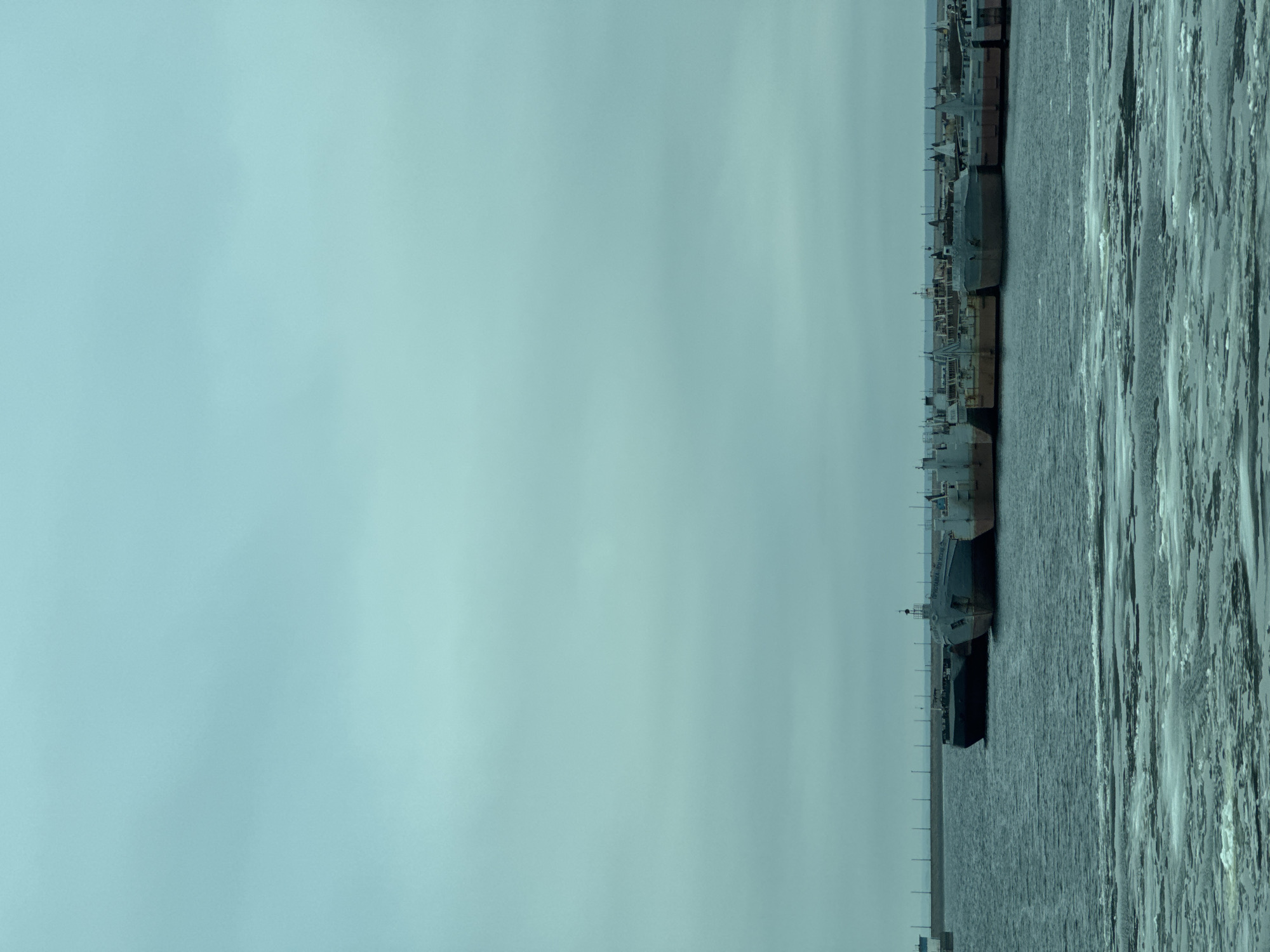 The image captures a broad, horizontal view of a winter or early spring scene along a body of water in Chaikovsky, Russia. The composition is divided horizontally: the upper two-thirds are dominated by a vast, overcast sky, while the lower third features water and ships.
On the left side of the frame, a continuous line of large, industrial ships, likely cargo vessels or barges, is docked along a riverbank or quay. The vessels are predominantly grey and dark-colored, appearing robust and utilitarian. They are moored side-by-side, extending into the distant haze. The water immediately adjacent to these ships is open and dark.
Towards the right side of the image, the water surface is extensively covered with fragmented ice floes, ranging from large chunks to smaller pieces, indicating cold weather. The ice creates a textured pattern of light and shadow across the water. A series of slender, vertical posts or bollards are visible along the edge of the water, separating the open water near the ships from the ice-covered expanse.
The sky is a uniform, light blue-grey, completely overcast, suggesting a dull, cloudy daytime. No sun is visible, and the light is soft and diffused. On the hull of one of the ships, approximately in the middle of the docked line, faint Cyrillic text can be discerned, reading "УК "МАЛАЯ ВОЛГА"". No people or discernible activity are visible in the scene, conveying a quiet, industrial setting.