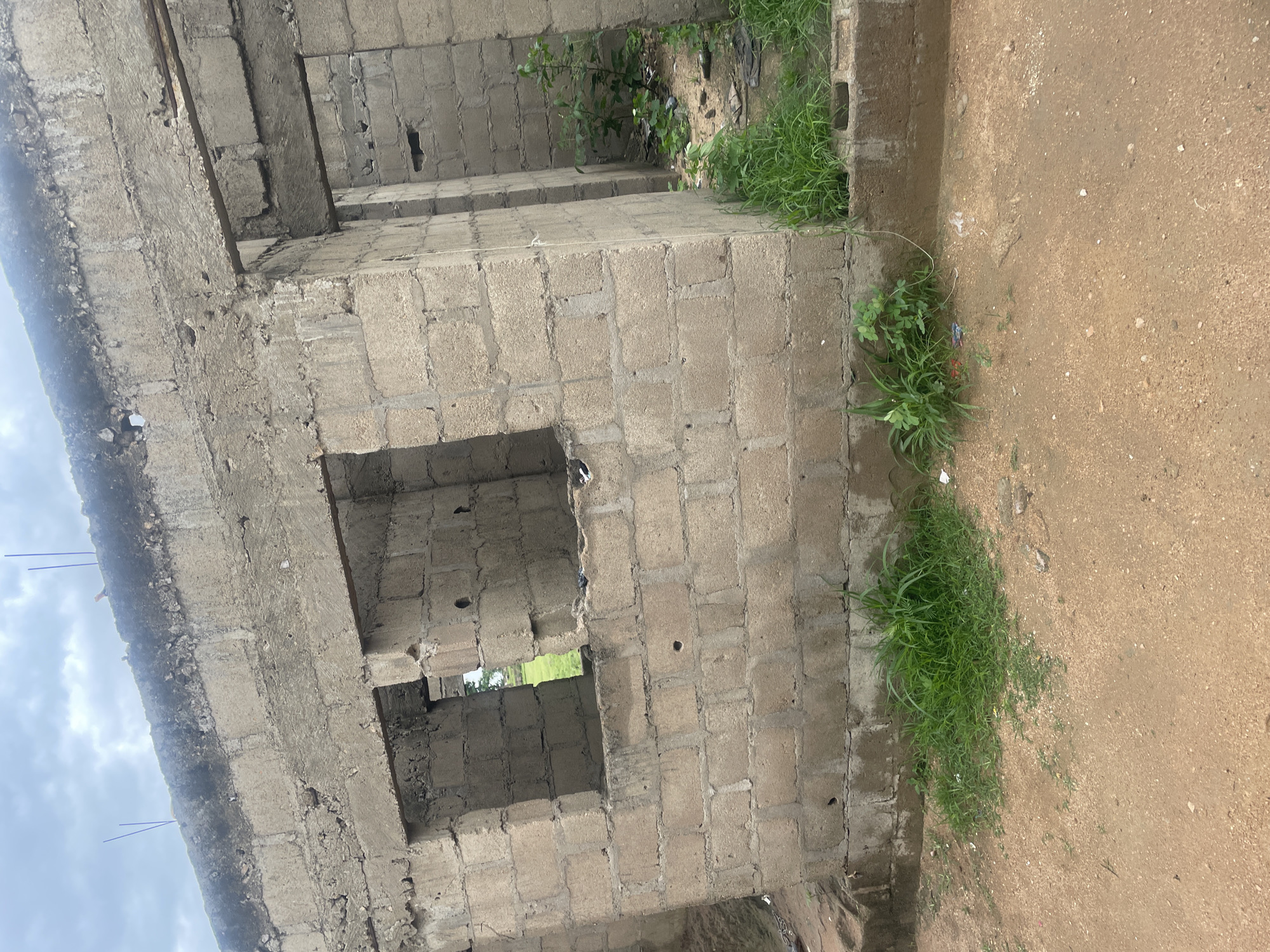 The image shows an unfinished building constructed from concrete blocks in Jalingo, Nigeria. The building has open window frames and doorways, revealing a glimpse of green grass and some debris inside. The structure appears to be in a state of disrepair or construction, with rough concrete edges and some exposed rebar at the top. The ground in front of the building is dirt and gravel, with patches of grass growing along the base. The sky above is cloudy. There are no people or animals visible in the image.
