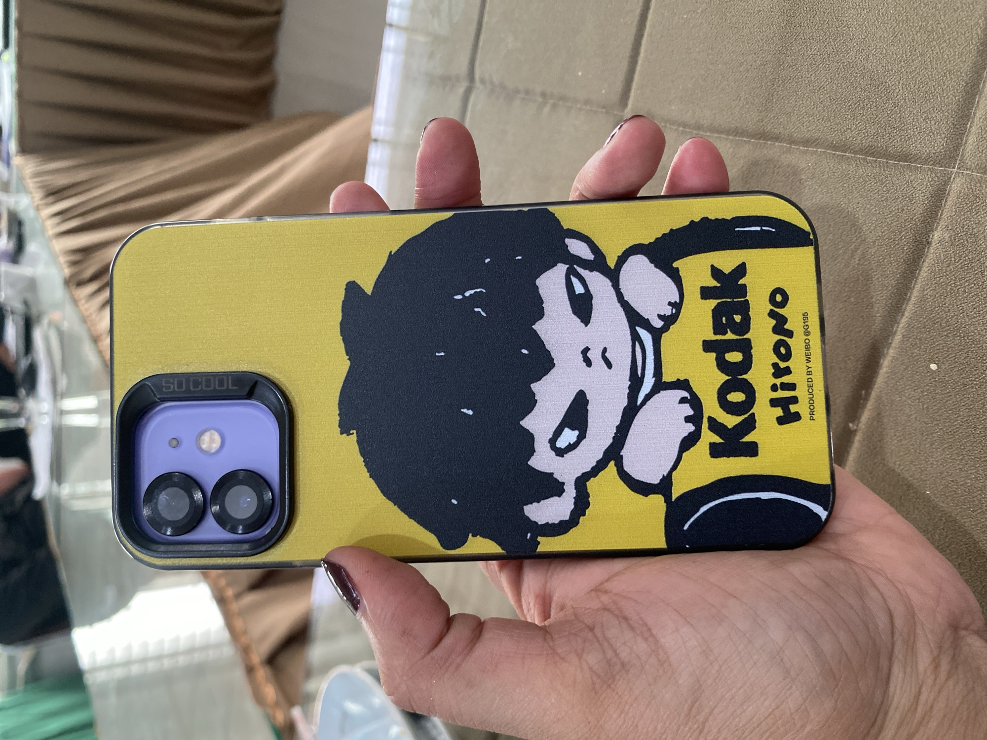 An iPhone case with a yellow background and a cartoon character is held by a person's left hand. The cartoon character has black hair and is dressed in a pink shirt. Below the character, the word "Kodak" is printed in black, followed by "Hirono" in a stylized font. At the bottom of the case, it says "PRODUCED BY WEIBO @G195". The phone has a dual-camera system with purple housing. The background is blurred, showing a table with a brown tablecloth and drapes. The lighting suggests an indoor setting. The city of Lhokseumawe is not depicted in this image.