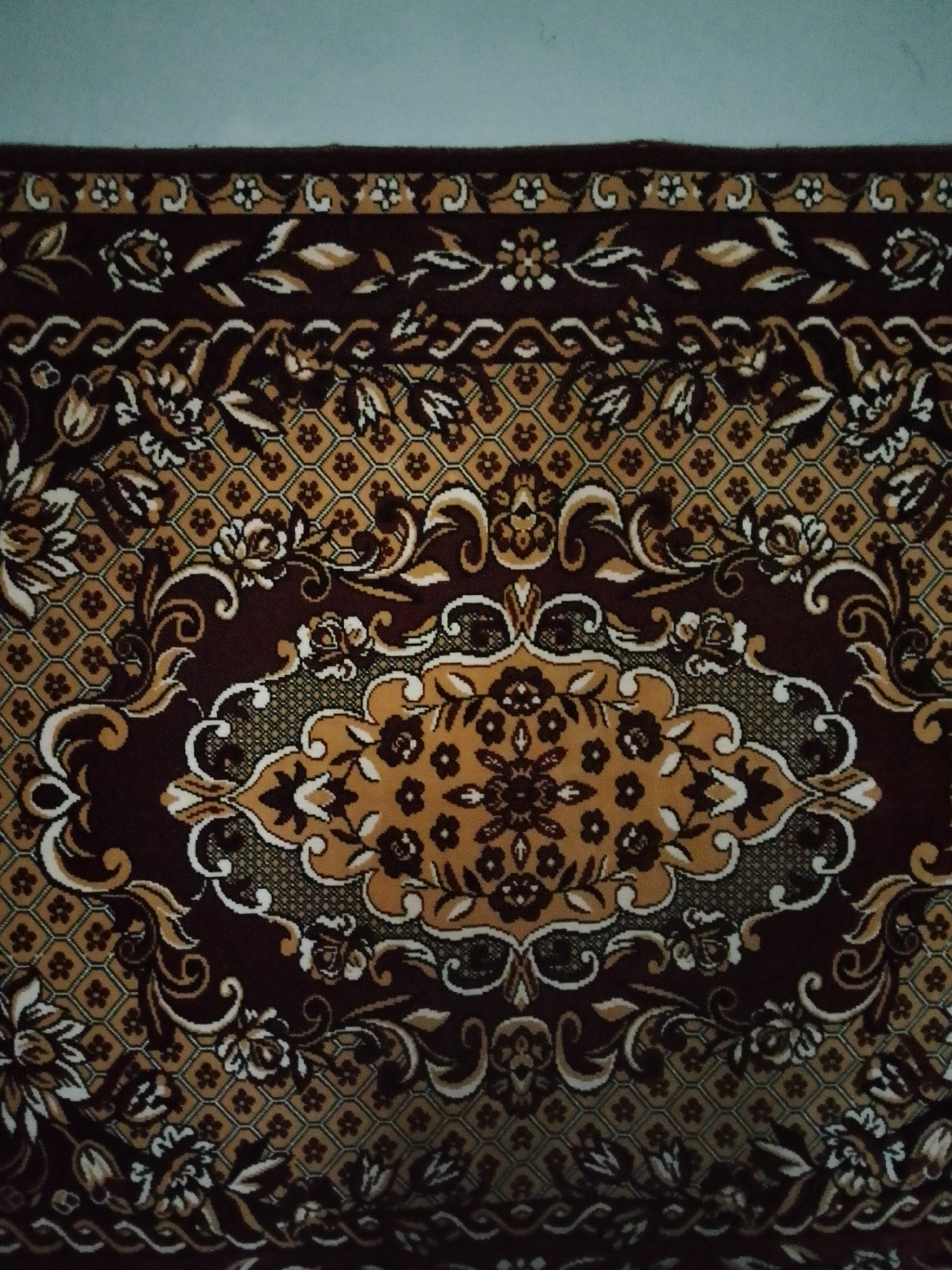 A rectangular textile, identified as a rug or carpet, is depicted from an overhead or slightly elevated perspective, positioned in an indoor setting in Unknown, Unknown. The rug features an ornate, symmetrical design dominated by a color palette of dark brown, gold, and cream. A large central medallion, gold with dark brown floral patterns and outlined in cream, forms the focal point. The main field of the rug is dark brown, adorned with scattered floral and foliate motifs in gold and cream, along with a repeating geometric lattice pattern of gold squares containing a small stylized cross or asterisk, set against a dark brown background. Multiple borders frame the rug, including bands of cream scrollwork, the geometric square pattern, and a wider border densely populated with stylized leaves and flowers in gold and cream on a dark brown ground. The outermost edge is a solid dark brown band. The top edge of the rug is positioned against a plain, light blue-grey surface, likely a wall or adjacent flooring. No individuals, actions, or interactions are visible in the scene.