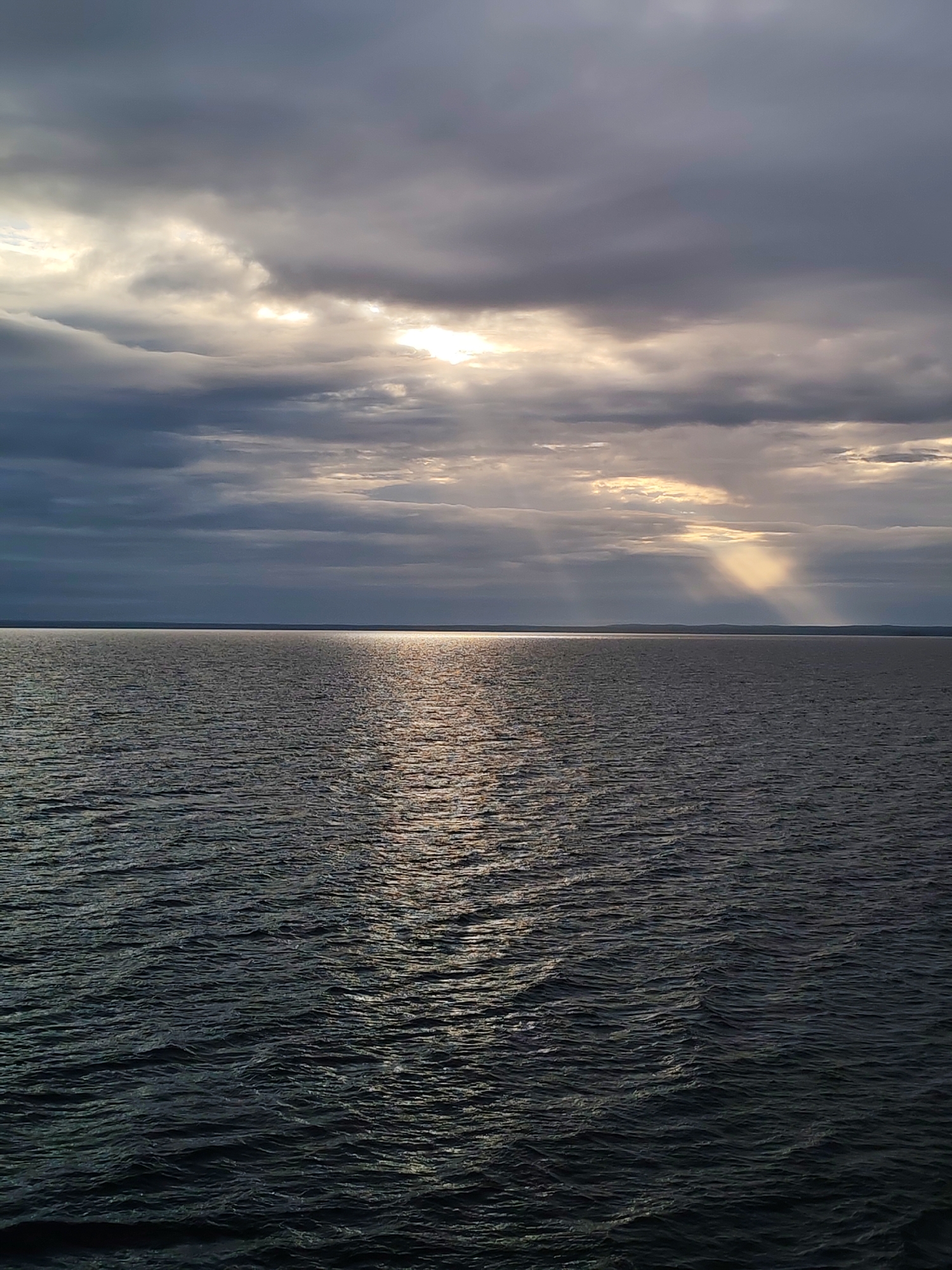 This media file presents a wide, open view of a vast body of water under a dramatic, cloudy sky. No people or artificial objects are visible, emphasizing the natural environment.
The scene is dominated by the dark, choppy water in the foreground and mid-ground, showing numerous small waves and ripples. A bright, shimmering path of sunlight reflects intensely on the water, stretching from the horizon towards the viewer.
Above the water, the sky is heavily overcast with dense, grey clouds. However, the sun is breaking through in several areas, creating brilliant streaks of light. Most notably, distinct crepuscular rays (sunbeams) are visible descending from the clouds on the right side of the image, illuminating parts of the water and horizon with a soft, ethereal glow.
A faint, dark landmass can be seen stretching across the distant horizon, suggesting the far shore of a large lake or sea. The quality of light, with the sun low and piercing through clouds, indicates either early morning or late afternoon (sunrise or sunset). The overall weather appears to be overcast with intermittent breaks of sunshine. The color palette is composed mainly of deep greys and blues for the water and clouds, contrasted by the bright white and golden hues of the sunlight.
The image conveys a sense of vastness, tranquility, and natural drama, possibly hinting at a momentary break in an otherwise cloudy day. No text is visible in the image. The location context is Kuzaranda, Russia.