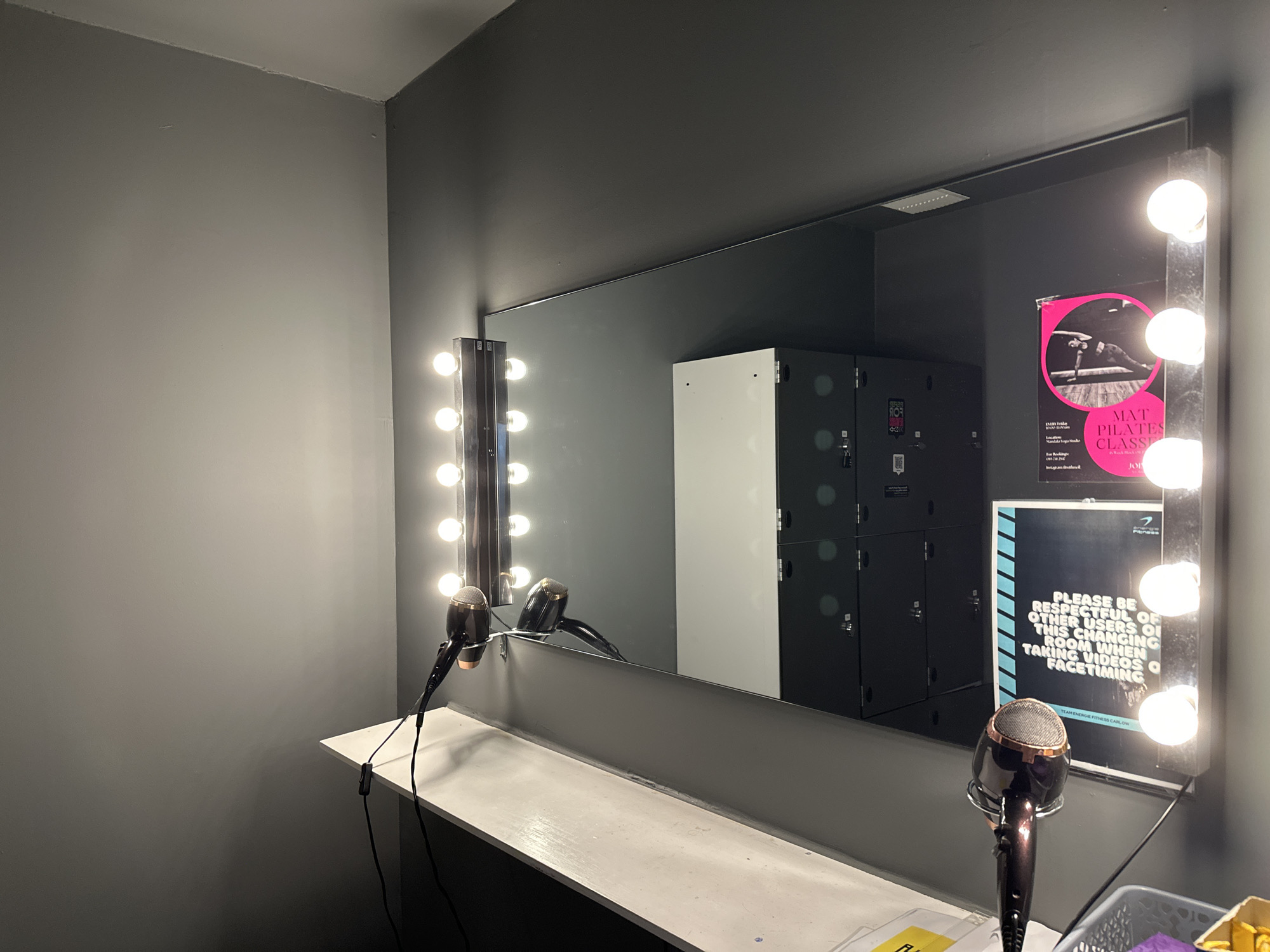 The image shows a changing room with a large mirror adorned with light bulbs, typical of a dressing room or vanity area. Two hairdryers are visible, one on the left side of the mirror and another in front of the mirror on a shelf. Behind the mirror, there are lockers, and on the right side of the mirror, there are two posters. One poster advertises "MAT PILATES CLASS" and the other has a message for users of the changing room regarding respectful behavior when taking videos. The room appears to be clean and well-maintained. The overall impression is of a functional changing room within a fitness or wellness facility.