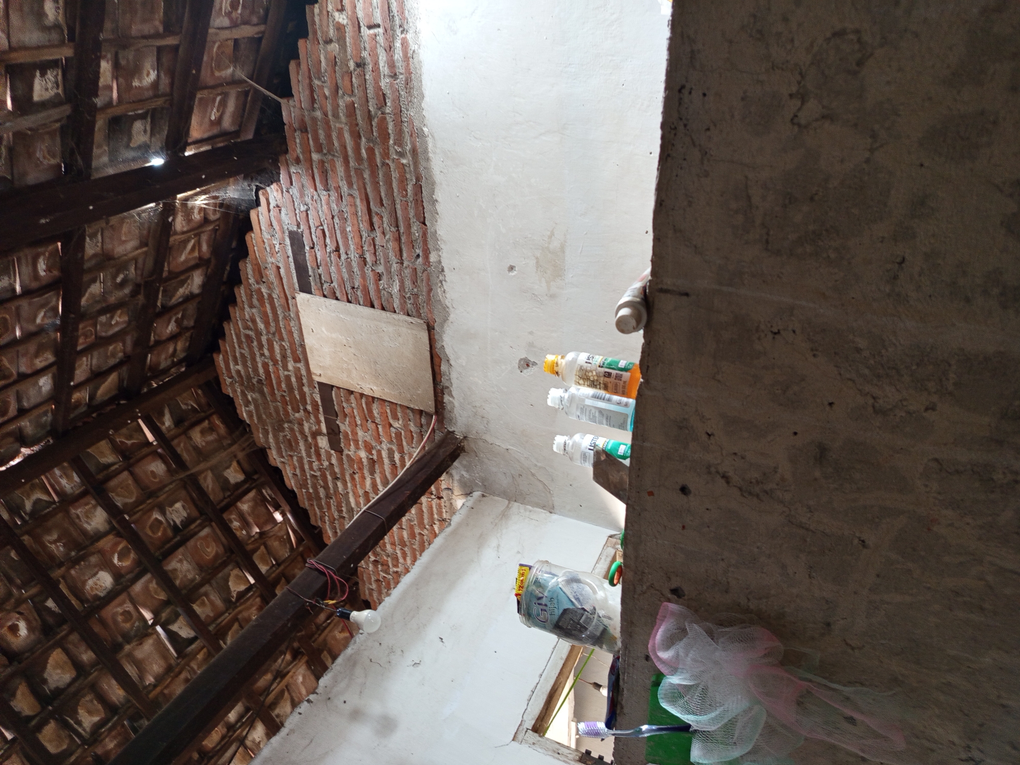 The image depicts an interior view of what appears to be an unfinished or partially constructed room with exposed brick walls and a tiled roof supported by wooden beams. A lightbulb hangs from a wire attached to one of the beams. In the foreground, a concrete ledge runs across the bottom of the frame. On this ledge, several items are placed, including a toothbrush, a jar with a label that reads "Giv Hijab," two clear plastic bottles with white and green labels, and another clear plastic bottle with an orange label. There are also what appear to be mesh bags or scrubbers to the left of the toothbrush. The walls are a mix of exposed red brick and white plaster. The roof is made of terracotta tiles supported by dark wooden beams. The lighting suggests it is daytime, with light filtering through gaps in the roof. The scene appears to be in a residential setting in Tebel, Indonesia.
