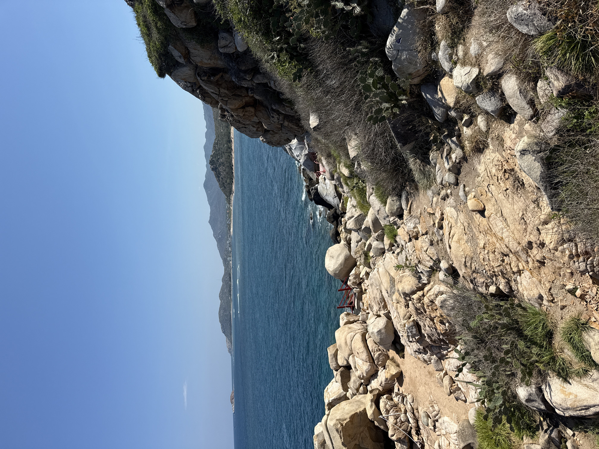 The image depicts a rocky coastline in Vinh Hai, Vietnam, seen from a high vantage point looking out over the ocean. The foreground features a steep slope covered in large boulders and sparse vegetation, including cacti and dry grasses. The ocean stretches out to the horizon, where distant mountains are visible. A red structure, possibly a platform or small bridge, is perched among the rocks near the water's edge. The sky is clear and blue.