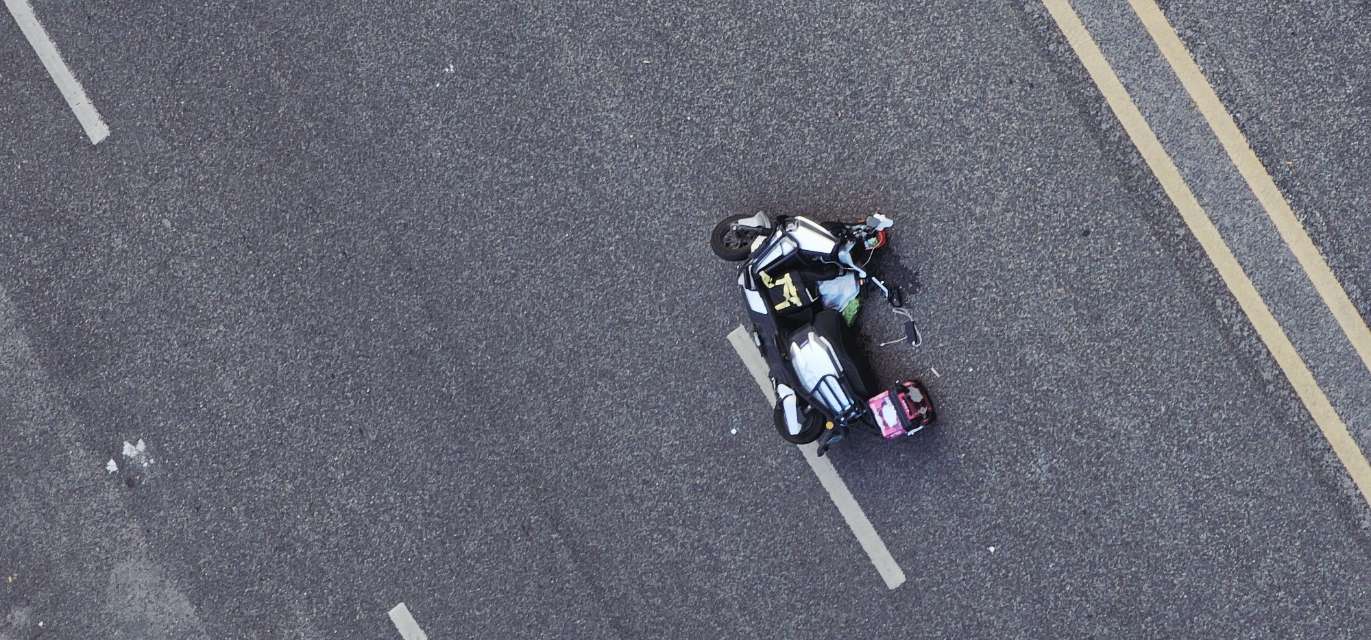 The image shows a fallen scooter lying on its side on an asphalt road in Jiulongpo District, China. The road markings include a double yellow line and white dashed lines. The scooter is white, black, and has a pink cargo box attached to the rear. Various items, possibly luggage or cargo, appear to be scattered around the scooter. The scene appears to be a traffic incident, with the scooter being the central subject. There is no visible presence of people.