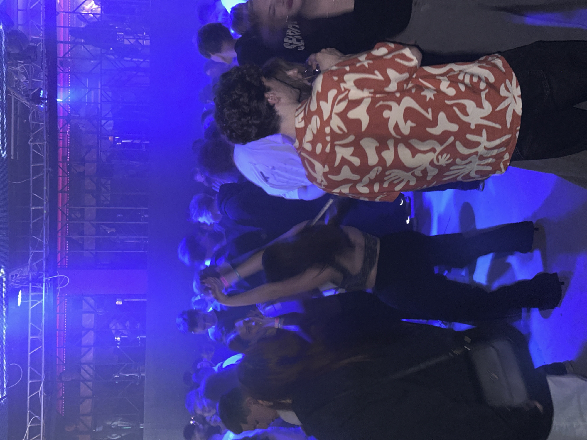 A crowd of people is gathered in a dimly lit, indoor space. Blue light floods the scene, illuminating faces and figures. In the foreground, a woman with long dark hair raises her arms. A man in a red and white patterned shirt stands near other individuals. Metal scaffolding is visible on the left side of the frame, suggesting a stage or performance area. Given the information provided, it is likely the location is a concert venue or nightclub in Izhevsk, Russia. The people appear to be enjoying a performance.