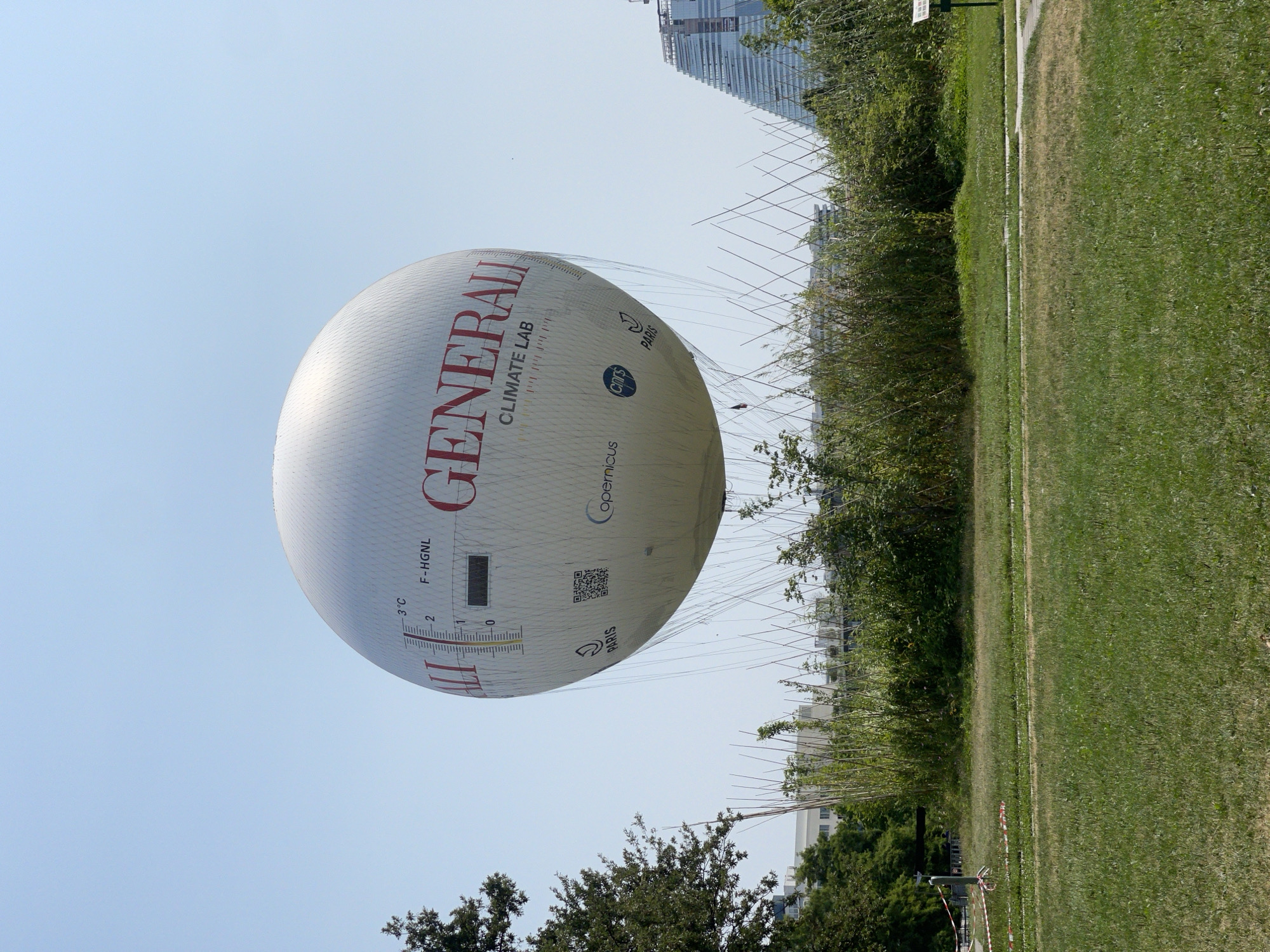 A large, white, spherical hot air balloon with the "GENERALI CLIMATE LAB" logo is tethered in a park-like area. The balloon has a temperature gauge marked with "3°C" and "0" at the bottom, as well as the text "F-HGNL". Various logos, including "PARIS", "Copernicus", and "cnrs", are visible on the balloon. In the background, there are buildings and trees, with a distinctive modern, stepped glass building visible on the right. The foreground consists of a grassy field with some pathways and safety tape. The sky is clear and blue. The scene appears to be an outdoor event or exhibition in Paris.