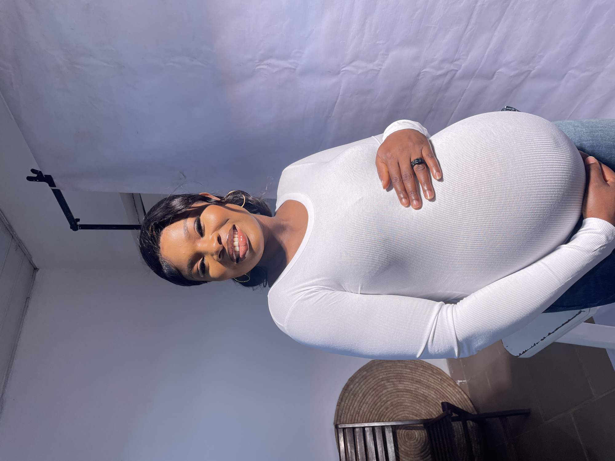 A pregnant woman stands smiling in what appears to be an indoor photo studio. She wears a white long-sleeved shirt and jeans and has her hands gently placed on her pregnant belly. The backdrop behind her is a large white sheet, possibly used for photography. To the left, there is a light stand. A chair sits just out of view towards the bottom right of the picture with a circular decoration on the floor to the left of the chair. While it's not explicitly stated, the photo is said to have taken in Jos, Nigeria.