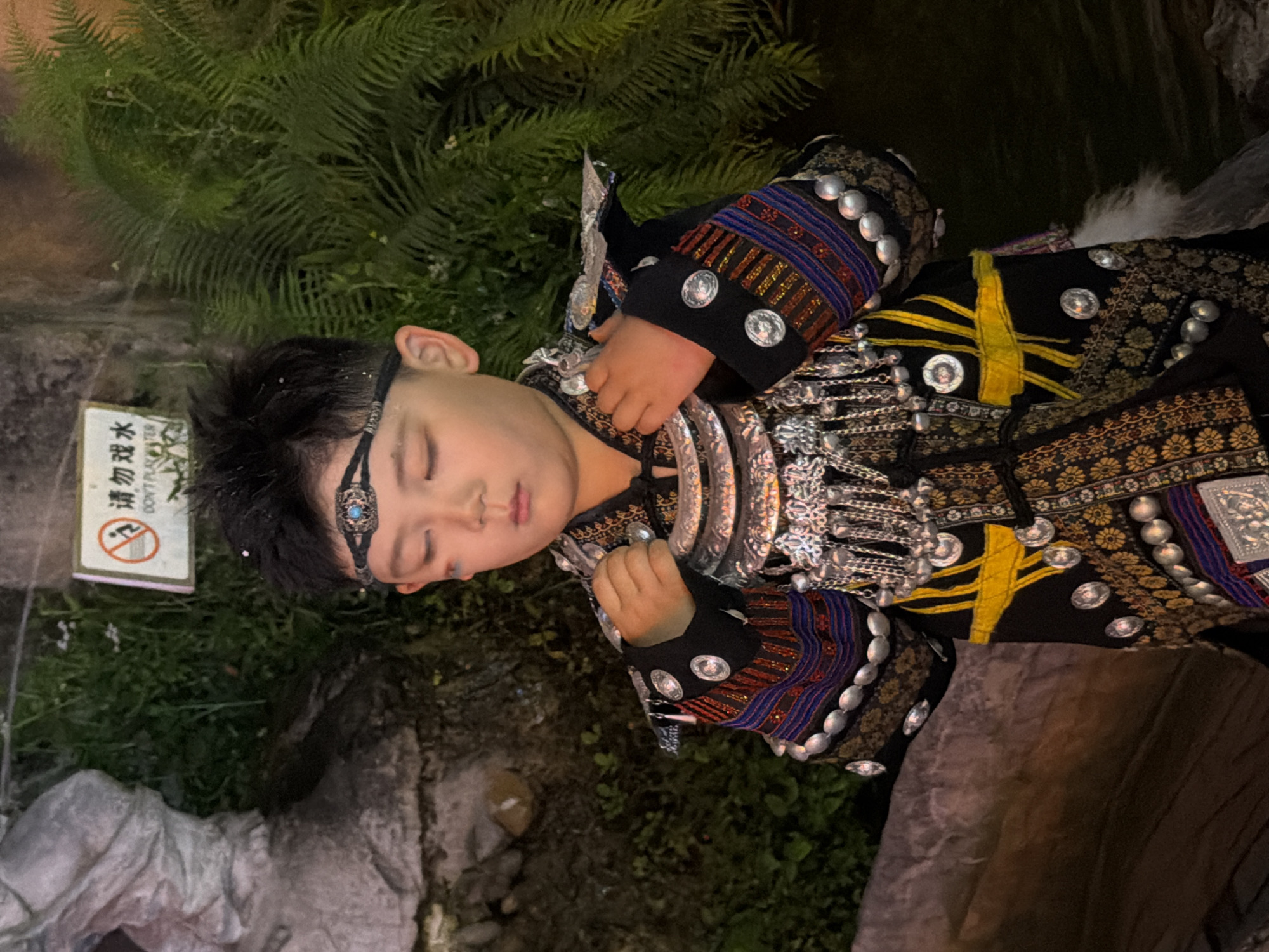 A young child, appearing to be of East Asian descent, is lying down, possibly asleep, and is dressed in elaborate traditional attire. The clothing is predominantly black with intricate patterns in gold, red, and blue. It is adorned with numerous silver circular ornaments and chain-like embellishments, particularly on the sleeves and chest area. A decorative headband with a blue jewel is worn across the forehead.
The child is positioned outdoors, with lush green foliage, possibly ferns, visible in the background. The ground appears to be rocky or uneven. To the left of the child, a white sign with Chinese characters and English text is visible. The text on the sign reads "请勿戏水" and "DON'T PLAY WATER". This suggests the location might be near a water feature where playing is prohibited.
The lighting suggests it might be evening or night, as the surroundings are dimly lit and there's a warm, artificial light illuminating parts of the scene. The overall mood is peaceful, with the child seemingly resting. The location is identified as Yuzhong District, China.