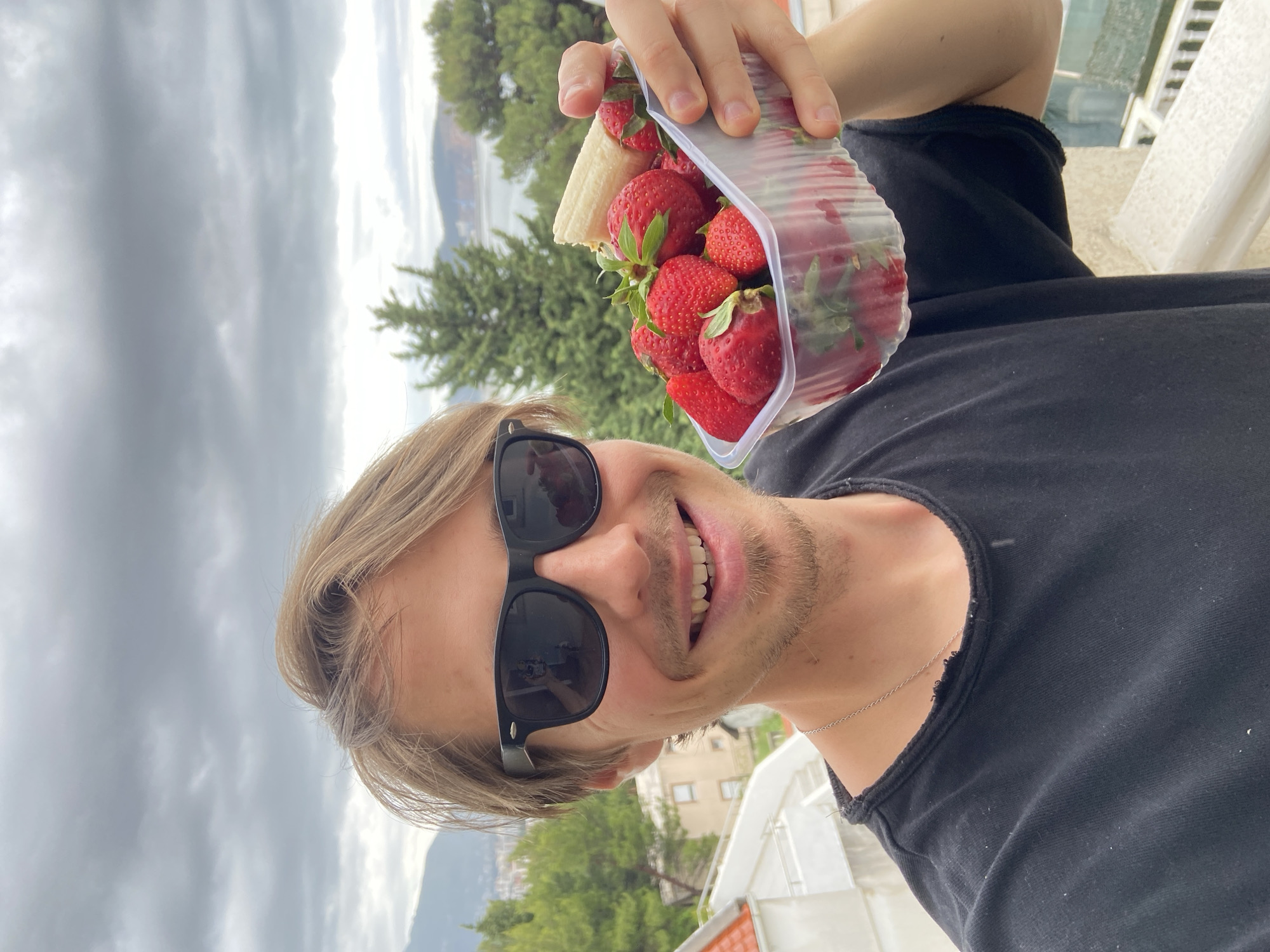 A man in black sunglasses and a black shirt holds a container of strawberries and a piece of white bread in front of him, smiling. The background suggests an outdoor setting, possibly a balcony, with trees, buildings, mountains, and a cloudy sky. The location appears to be Šušanj, Montenegro.