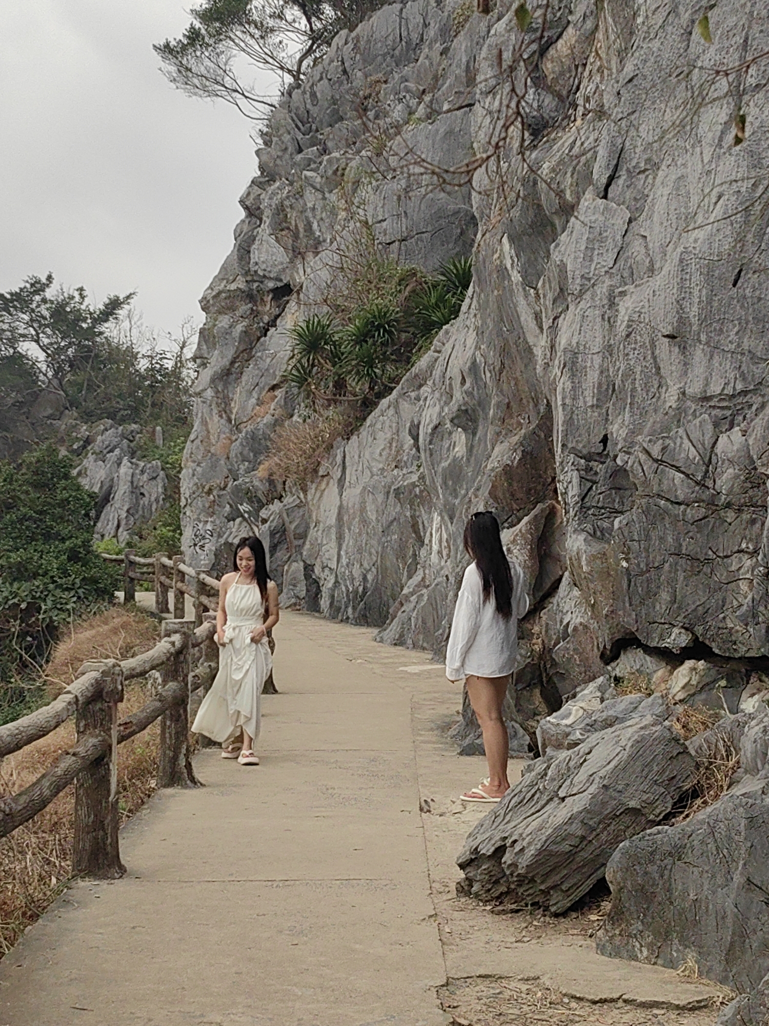 Two women are on a concrete path alongside a large, rocky cliff. The path is bordered on one side by a rustic, wooden fence. The woman on the left is wearing a flowing white dress and smiling. The woman on the right wears a white shirt and appears to be observing the cliff face. The environment includes trees, vegetation clinging to the rocks, and an overcast sky. The location is in Hải Phòng, Vietnam.