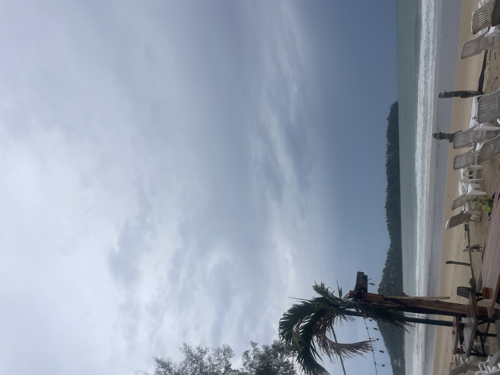 The image shows a beach scene in Choeng Thale, Thailand. The sky is overcast with white and grey clouds. The ocean is visible, with waves breaking on the sandy shore. A line of vegetation is present between the beach and the sky. In the foreground, there is a palm tree and other trees, as well as beach chairs and wooden tables.