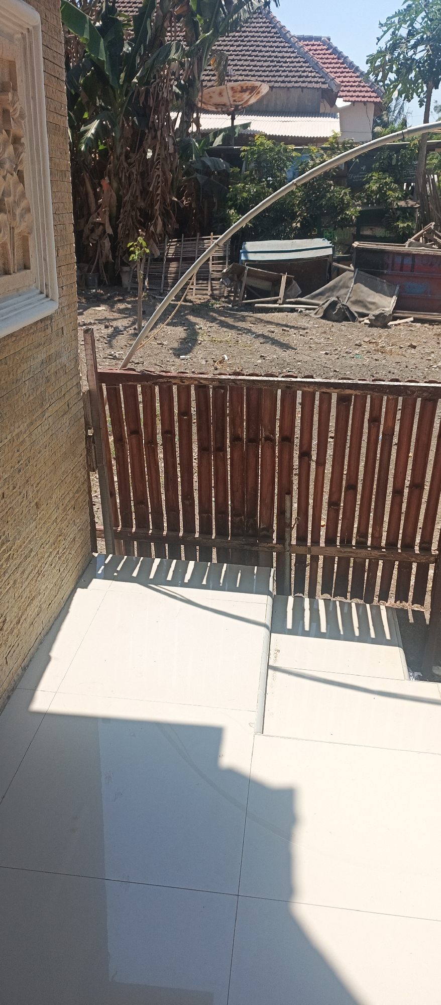 A daytime, eye-level, outdoor shot captures a close-up of a weathered bamboo fence in Jatisari, Indonesia. The fence, constructed from vertical bamboo stalks, is positioned in front of a tiled patio area. The patio itself is partially shaded by the fence, creating a pattern of light and shadow. Beyond the fence, a rustic scene unfolds with a satellite dish atop a house, banana trees, and various discarded objects including a boat, wooden crates, and tarps, suggesting a storage or work area. The overall environment appears to be a residential backyard or a similar outdoor space.