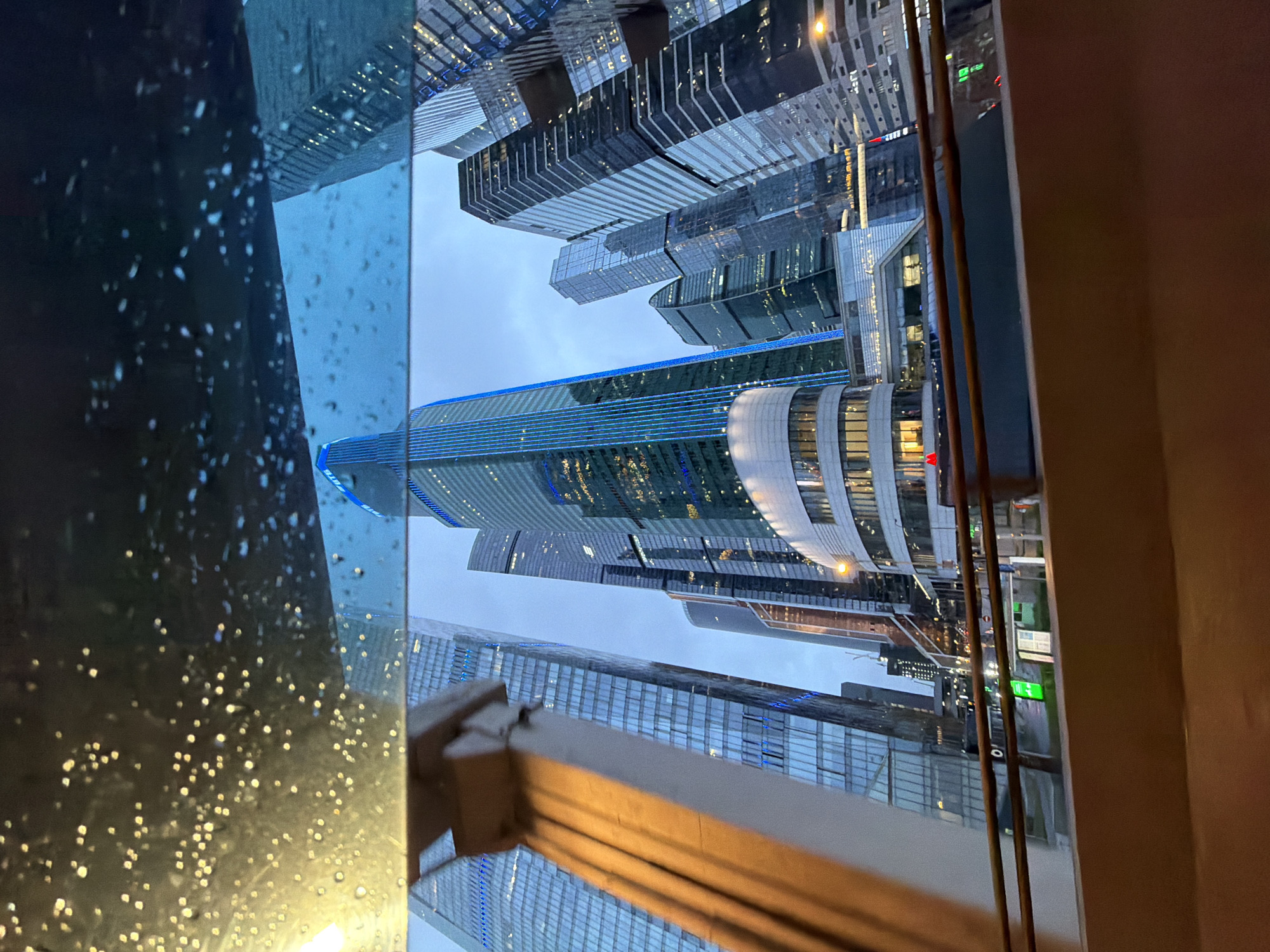 The image depicts a view of the Moscow International Business Center (Moscow City) through a window on a rainy day. The window pane is visible on the left side, covered with raindrops. The Moscow City skyscrapers, including the Evolution Tower, are prominent in the background. The buildings feature a mix of glass and steel architecture. Streetlights and other urban infrastructure are visible in the lower portion of the view.