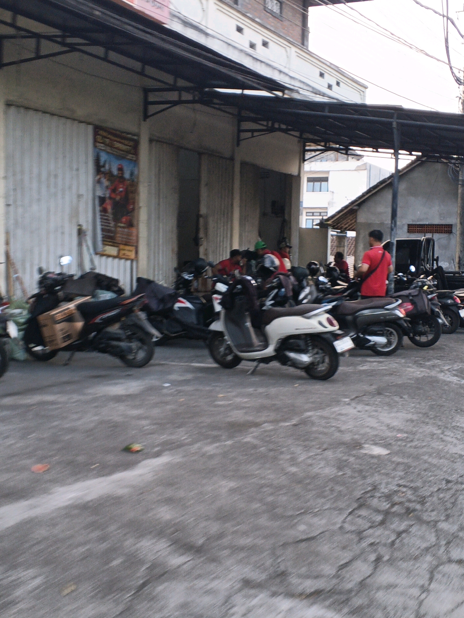 In Denpasar, Indonesia, a street scene unfolds outside a commercial establishment with a corrugated metal facade. Several motorcycles are parked on the cracked asphalt pavement. In the foreground, a light-colored scooter is parked facing left, with a dark-colored scooter to its right. Behind these, more scooters are visible, some with riders. One person, wearing a red shirt, stands near a parked motorcycle, facing away from the camera. The background features the building with its industrial-style shuttered entrances and a black awning extending over the front. A poster is affixed to the wall, depicting a person in red. The sky is bright and overcast.