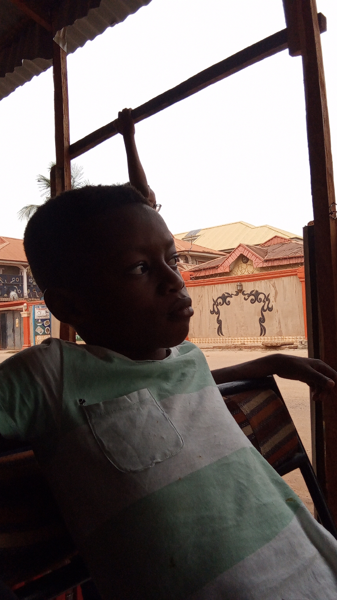 A young child, likely a boy, is depicted in a shaded, open-air setting, looking intently towards the right of the frame. His face, partially obscured by shadow, shows a contemplative or observant expression. He is wearing a striped t-shirt with alternating light green and white bands, featuring a small pocket on the left chest. His right arm rests on the back of what appears to be a patterned or striped chair.
Above him, another person's arm and hand are visible, holding onto a sturdy wooden beam that forms part of the overhead structure. A corrugated metal roof is partially visible in the upper left corner, suggesting a shelter or canopy.
The background is bathed in bright daylight, with an almost blown-out sky dominating the upper portion. Below the sky, several buildings with orange-brown tiled roofs and painted walls can be seen. A distinctive, light-colored wall with dark, elaborate decorative patterns resembling an arch is prominent in the middle distance. Sparse, palm-like foliage is also visible, silhouetted against the bright sky. The strong backlighting creates a stark contrast between the shaded foreground subjects and the brightly illuminated background. The scene suggests an outdoor, possibly urban or semi-urban environment in Sabon Tasha, Nigeria, during the day. No legible text is visible.