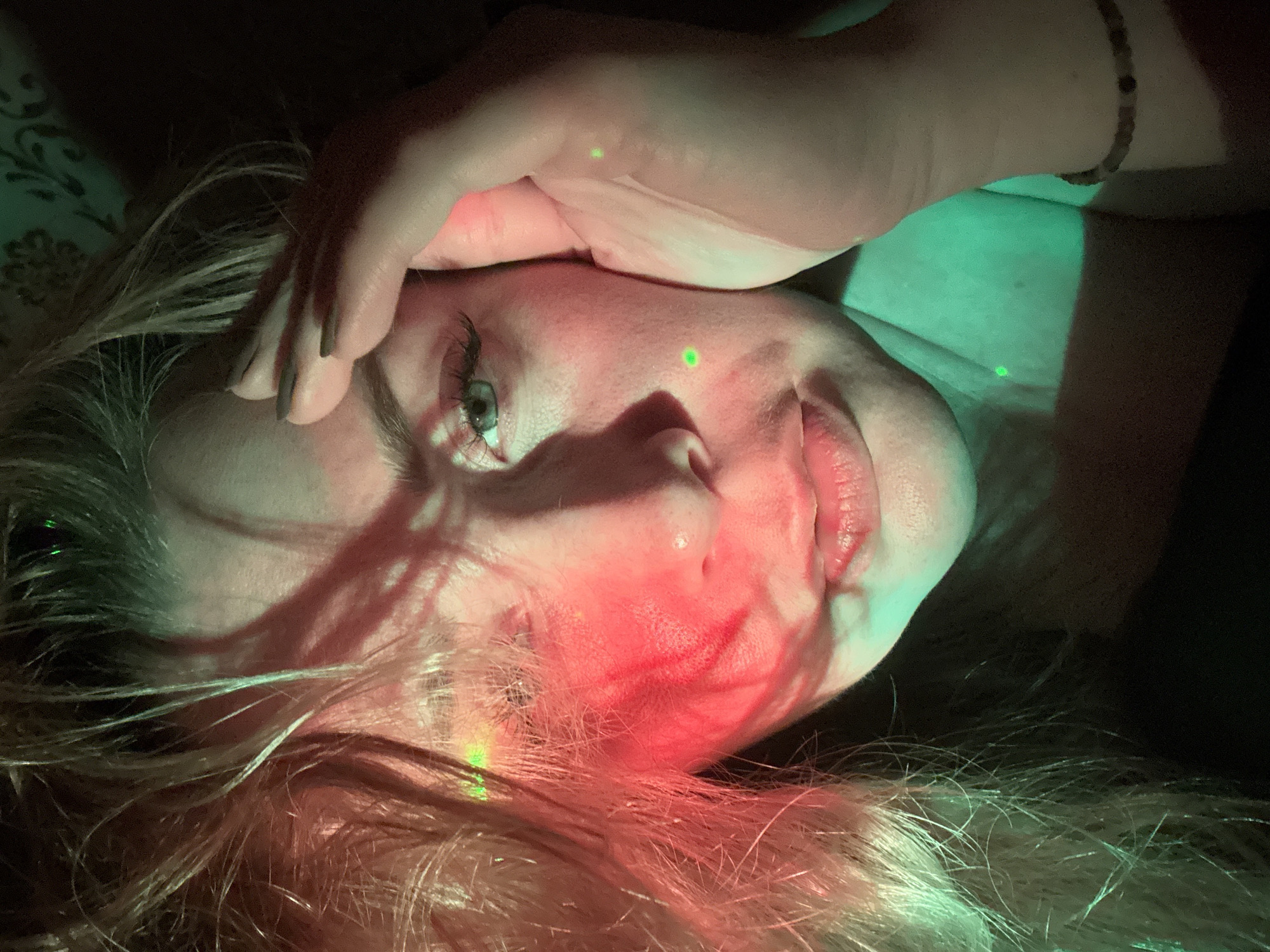 An adult female is depicted in a close-up portrait, located within the city of Veliky Novgorod, Russia. She has light-colored, disheveled hair partially obscuring her face. Her right side (viewer's left) is bathed in intense red light, illuminating her hair and the contours of her cheek and nose. Her left side (viewer's right) is illuminated by a strong green light, highlighting her left eye, cheek, and neck. Her left eye, with a light-colored iris (likely green or blue) and prominent eyelashes, is clearly visible, while her right eye is largely in shadow. Her lips are slightly parted. Several small, bright green laser dots are projected onto her skin; one is visible on her left cheek (viewer's right), another near her right temple (viewer's left), and a third on the back of her left hand (viewer's right). Her right hand (viewer's left), with dark-painted fingernails, is raised and positioned against her right temple/forehead. A beaded bracelet is worn on her left wrist (viewer's right). She is wearing a dark-colored garment visible around her neck and shoulders. The background is dark, featuring a faint, lighter-toned floral or damask pattern on the wall behind her. The overall scene is characterized by distinct, colored artificial lighting against a dark environment.