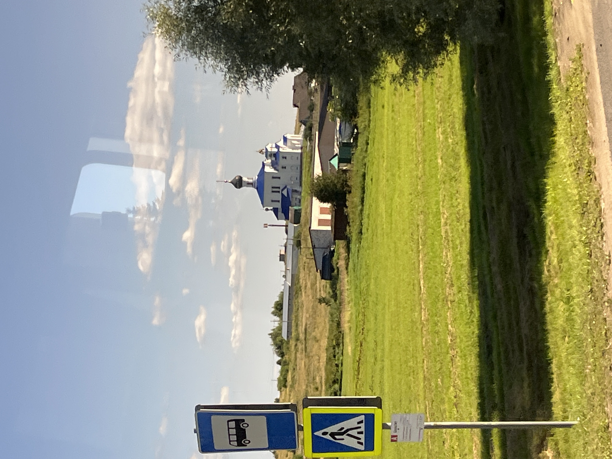 The image shows a rural landscape in Myadzyel, Belarus, on a sunny day with scattered clouds. In the foreground, a metal pole supports two traffic signs: a blue sign with a white bus icon indicating a bus stop, and a yellow sign with a black pedestrian icon indicating a crosswalk. Below these signs is a smaller white sign with text and a red letter "A".
Behind the signs, a vast, vibrant green field stretches towards a gently sloping hill. The field appears to be a mown meadow, with visible lines from the mowing equipment.
On the hillside, several buildings are visible. Prominently featured is a white Orthodox church with a blue dome, topped with a cross. Adjacent to the church are smaller buildings, some with brown or green roofs. Further in the background, a row of longer, lighter-colored buildings with industrial-style roofs are situated, suggesting a possible agricultural or commercial complex.
To the right of the frame, a large, leafy green tree partially obscures the view of the distant landscape. The sky is a clear blue, with soft white clouds scattered across it. Reflections of the sky and possibly trees or buildings are visible on what appears to be a window, suggesting the photo was taken from inside a vehicle. The overall scene depicts a tranquil, rural setting in the vicinity of Myadzyel.